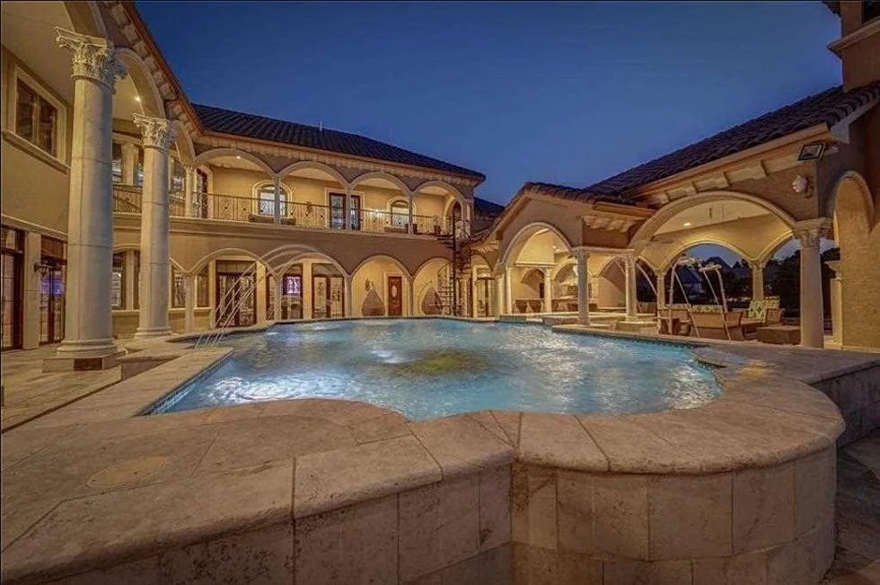 Luxurious mansion with illuminated swimming pool at dusk, surrounded by a two-story building with arched walkways and columns.