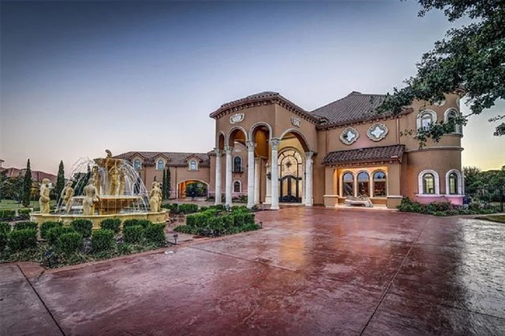 Large mansion with a decorative fountain in front, arched windows, and a driveway leading up to the entrance amid well-maintained landscaping.