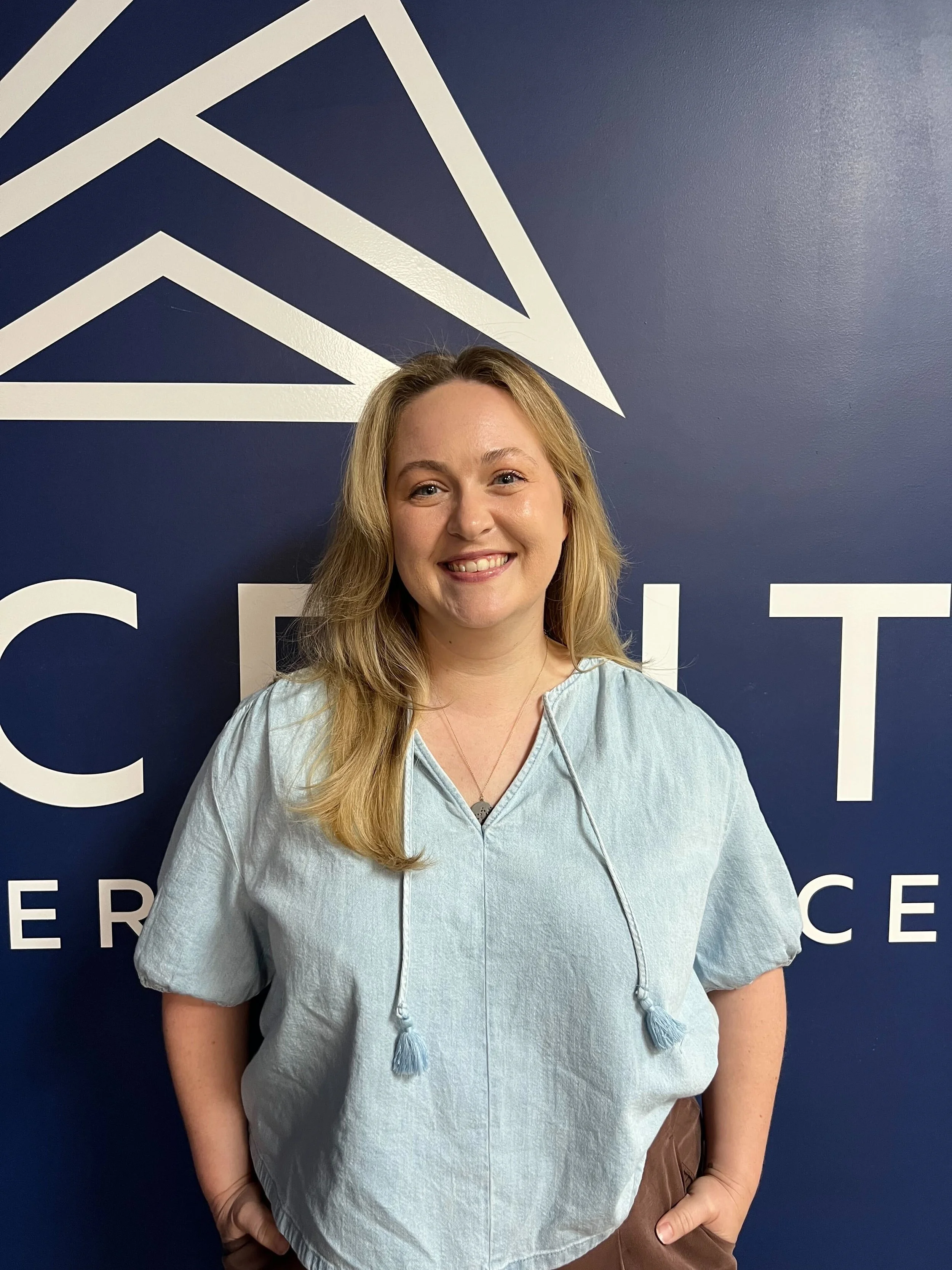 A young woman with blonde hair and blue eyes smiling in front of a blue wall with white geometric design and text.