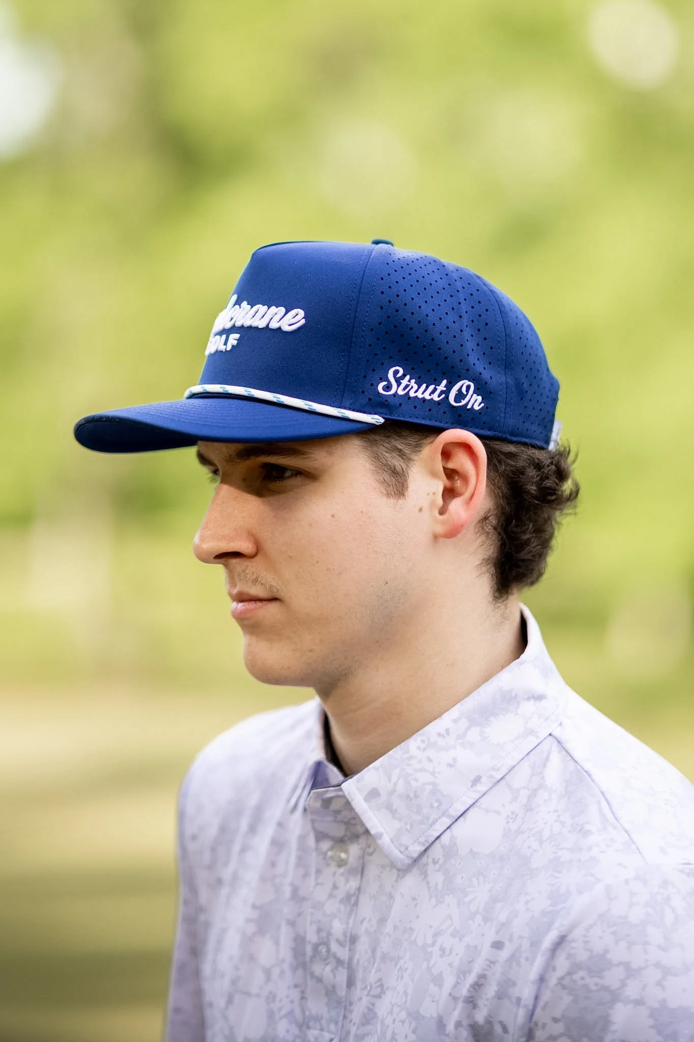A man wearing a white golf cap with 'Sandcrane Golf' logo stands outdoors with a grassy area in the background.