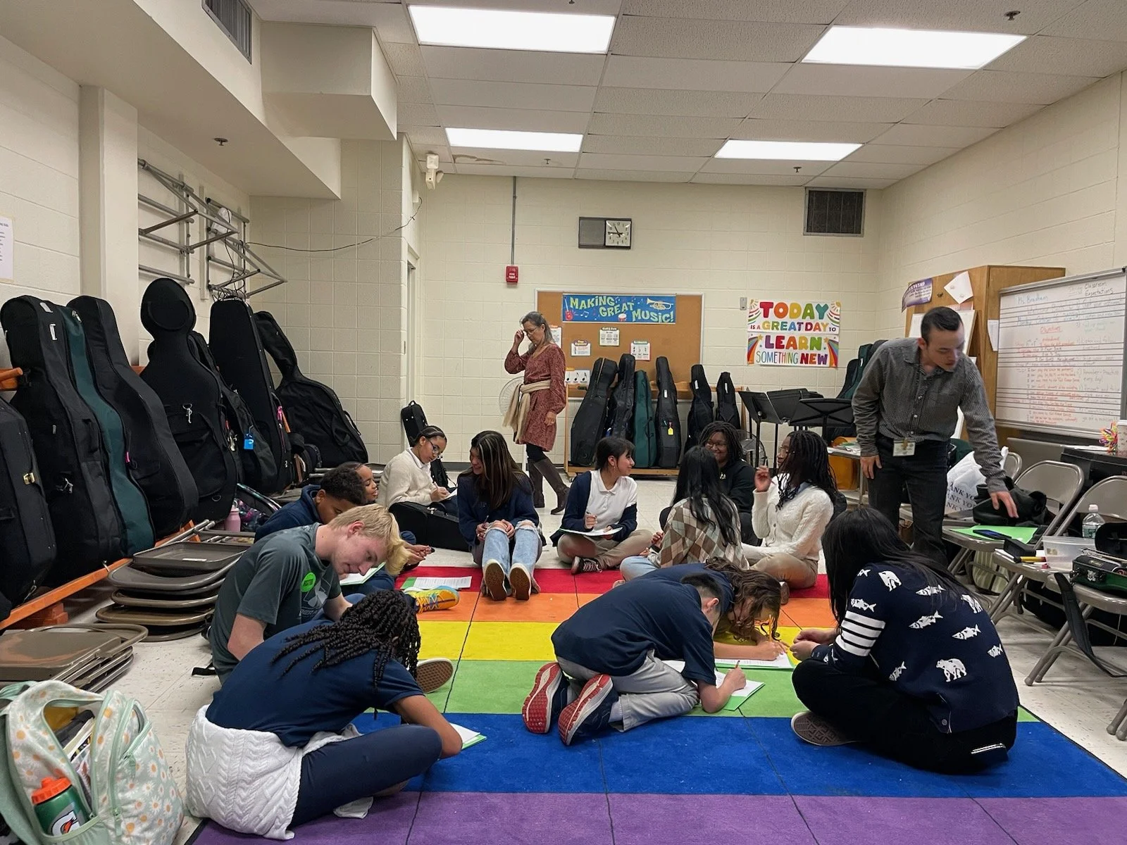 A classroom with students sitting on a colorful rainbow rug, some writing on paper, and a teacher standing nearby. Back wall has musical instrument cases, colorful posters, and a whiteboard.