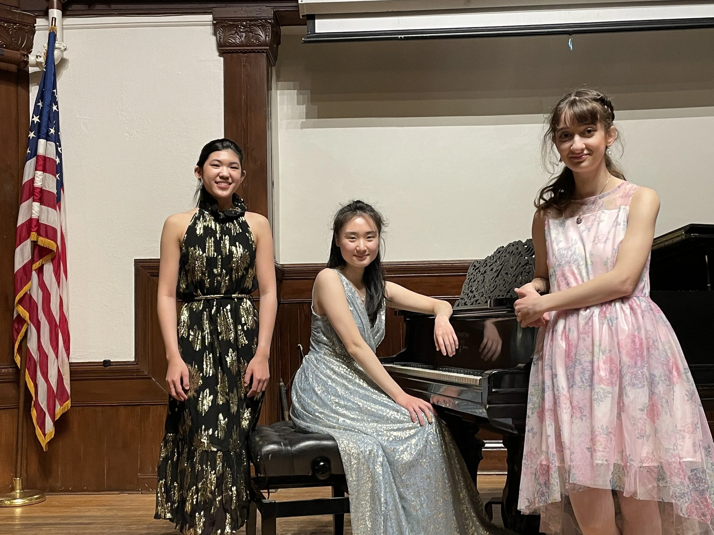 Three young women in elegant dresses posing around a black grand piano in a room with wooden paneling, an American flag, and a projector screen.
