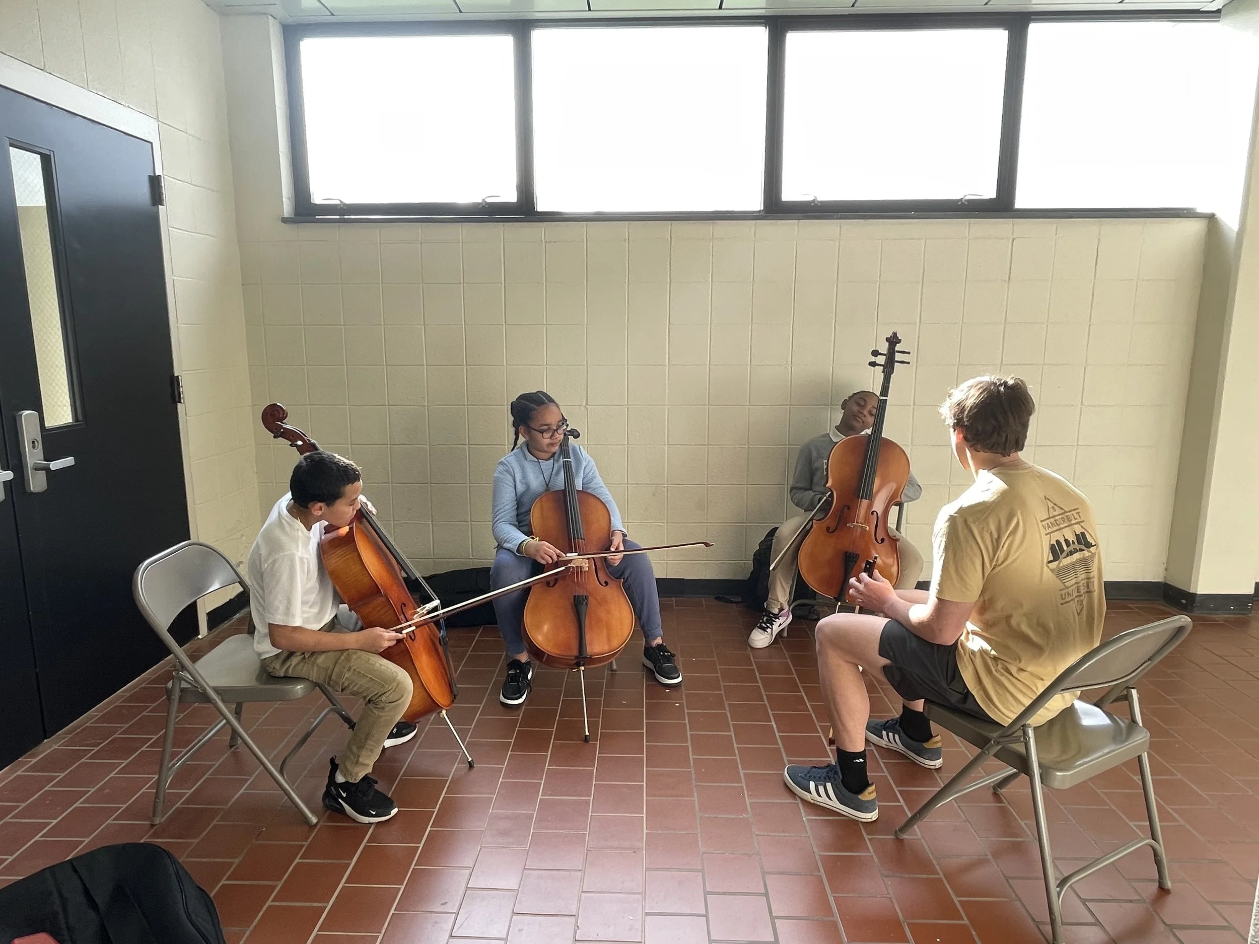Group of four children sitting in a circle in a music classroom, practicing with musical instruments. They are playing cellos and violin, with a music teacher guiding them.