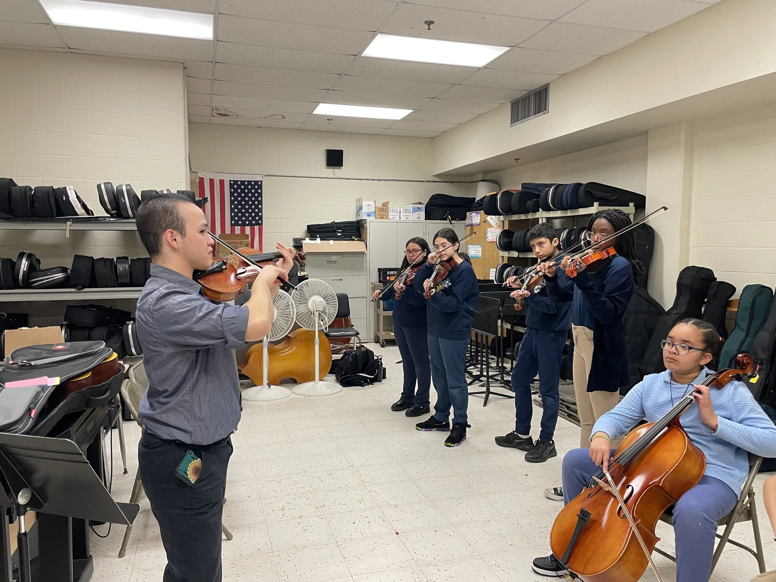 A music classroom where a group of young students are practicing string instruments, including violins and a cello, with an instructor.
