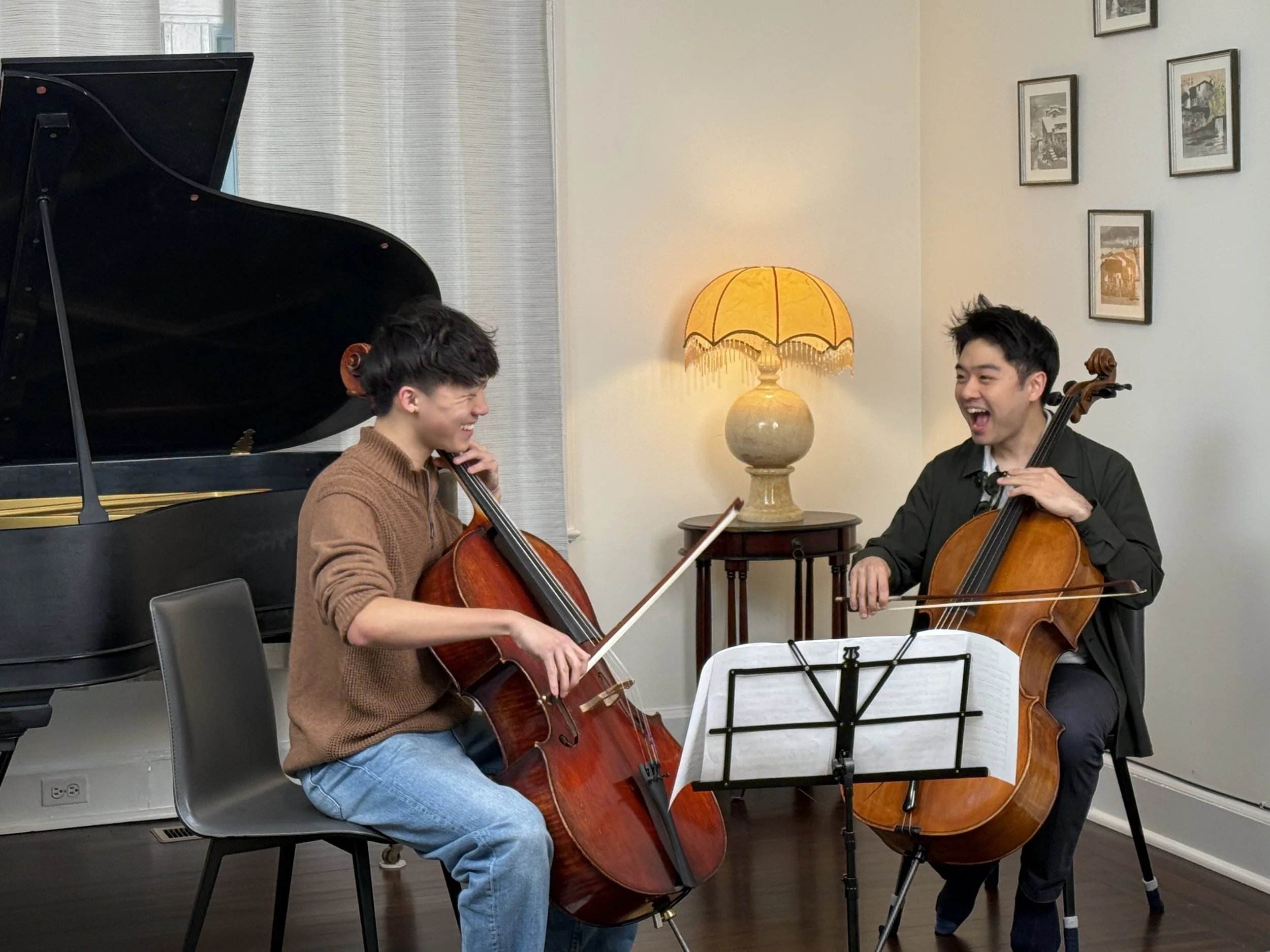 Two young adults playing cellos and smiling during a music session in a room with framed pictures, a table with a lamp, and a grand piano.