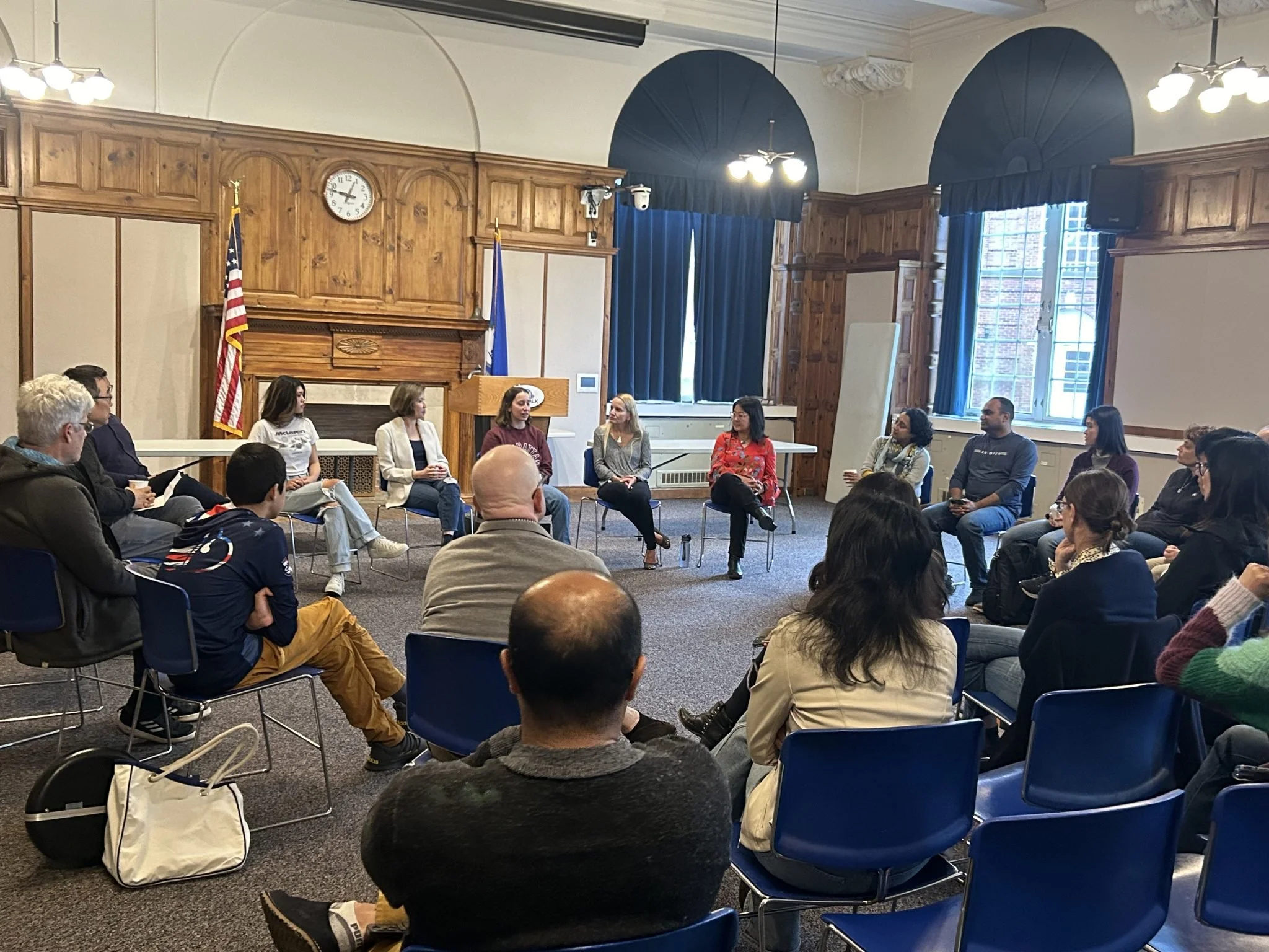 A diverse group of people sitting in a circle in a large, wood-paneled room, engaged in a discussion or panel. There are flags, large windows with blue curtains, and a fireplace on the wall.