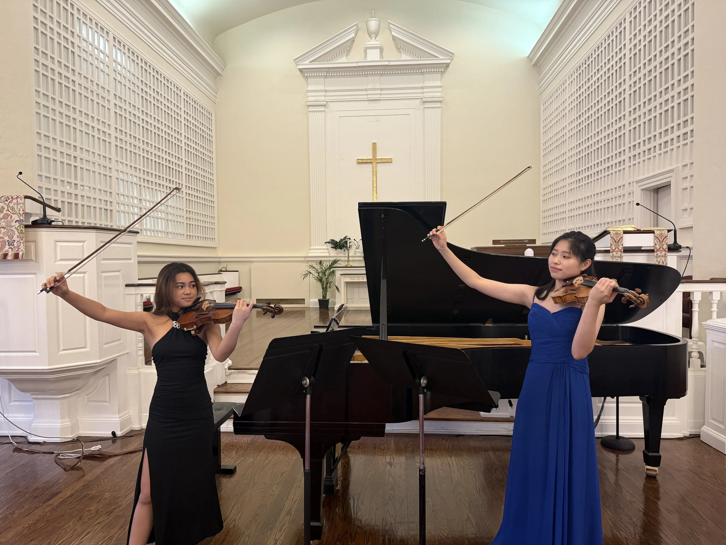 Two young women in formal dresses playing violins on stage in a church setting, with a grand piano, microphone stands, and a cross on the back wall.