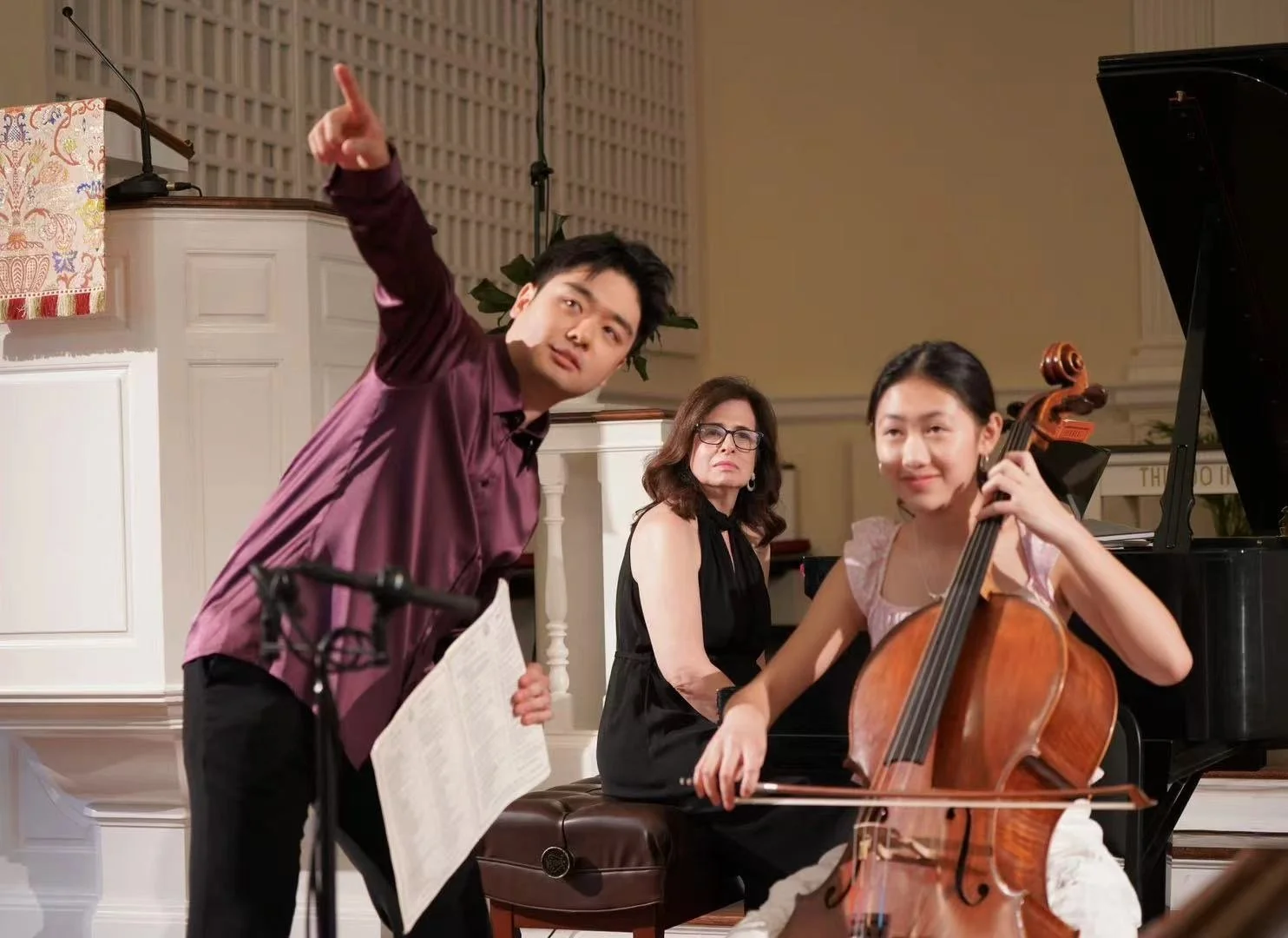 A man in a maroon shirt points upwards while holding sheet music, with a woman playing the cello and another woman sitting at the piano in a concert hall.