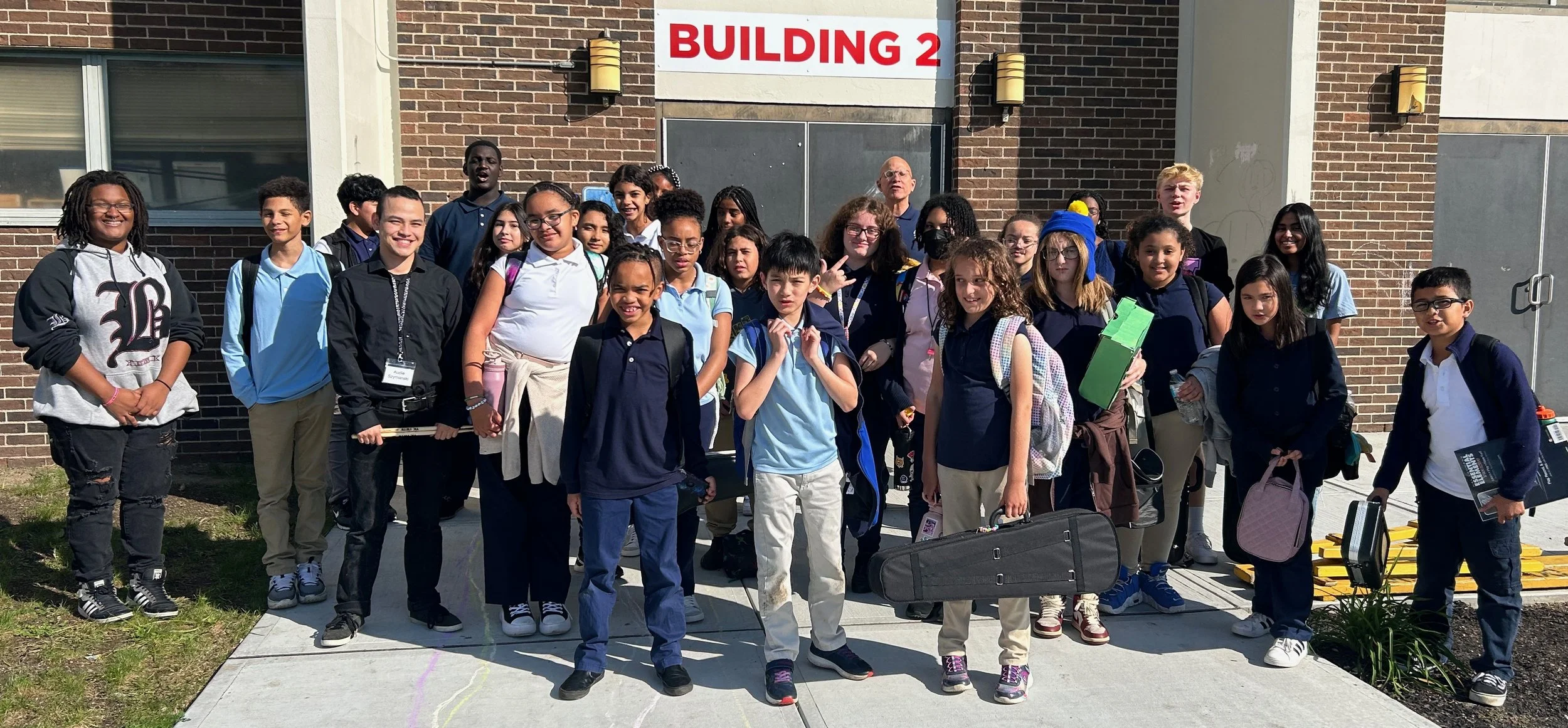 Group of middle school students and a teacher standing outside in front of a brick building labeled "Building 2". The students are holding backpacks and school supplies, and some are smiling at the camera.