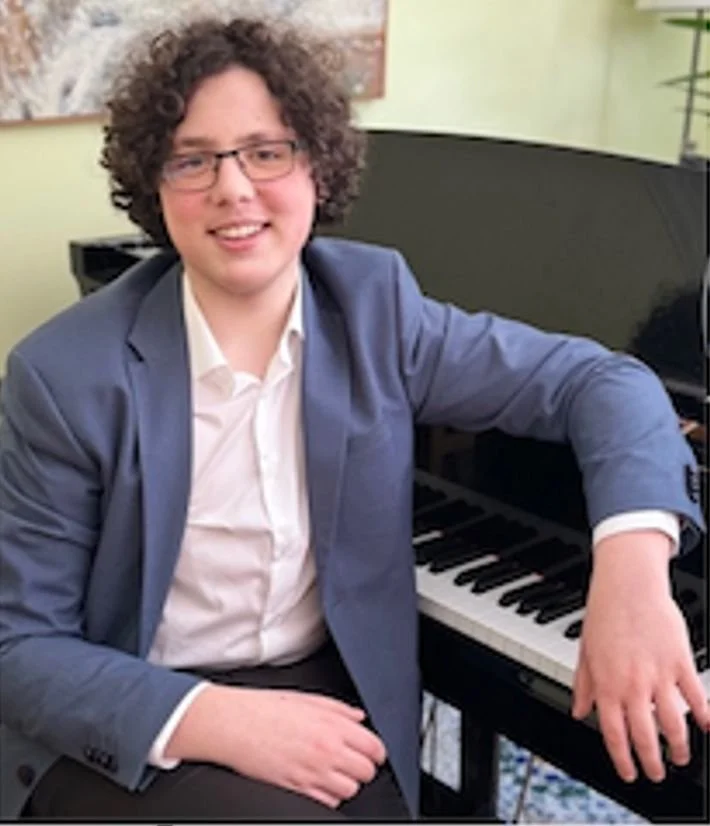 Man with curly hair and glasses sitting at a piano, wearing a blue blazer and white shirt, smiling indoors.