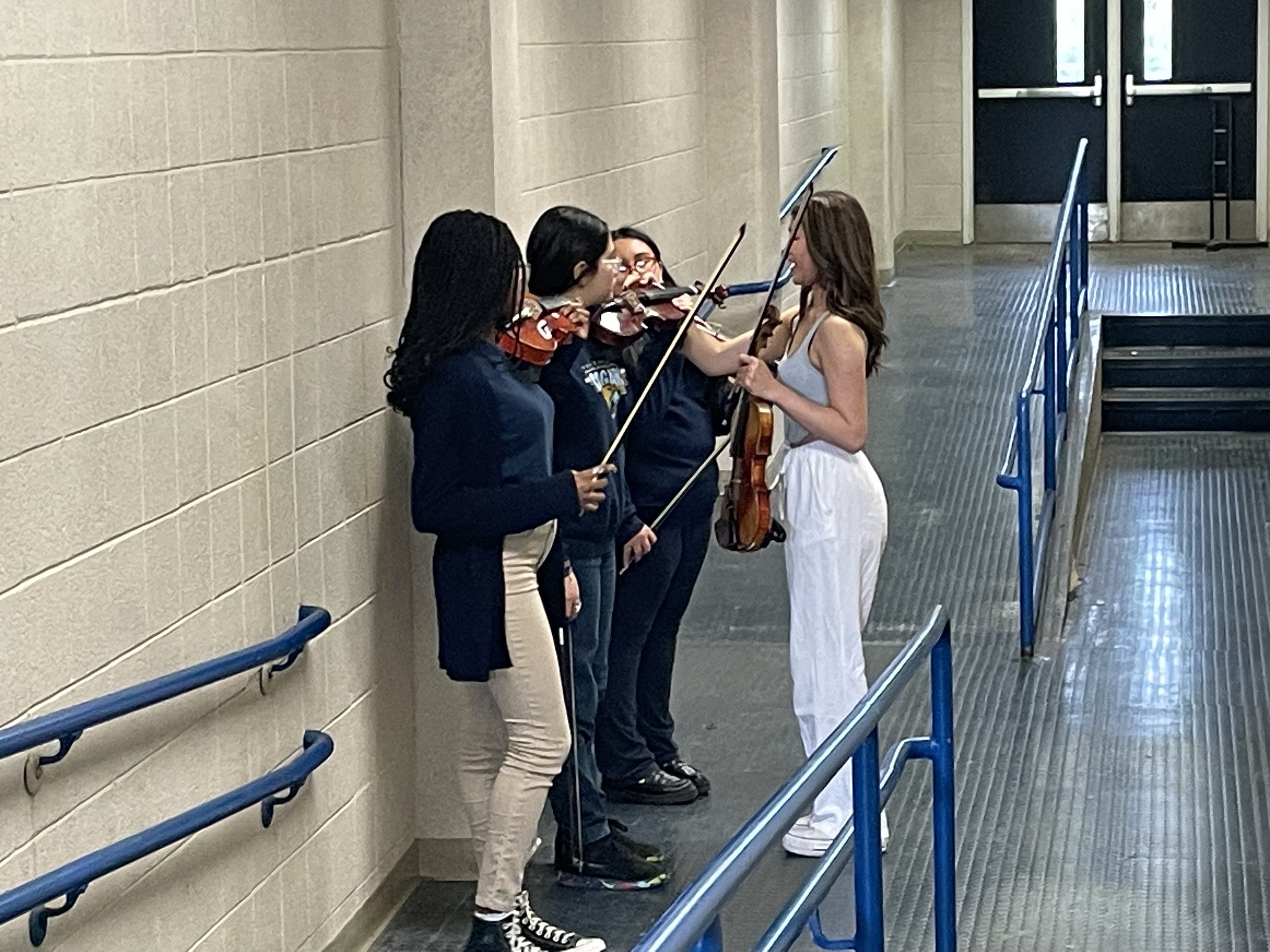 Four young women are standing against a beige wall, with two of them playing violins and the third speaking to the woman in white pants who is playing the viola. The women are engaged in a music practice or rehearsal in a corridor near a door with gl