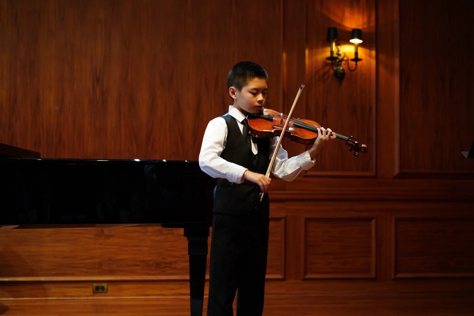 Young boy in formal attire playing violin on stage with a grand piano in the background, wooden paneling and wall sconces lighting.