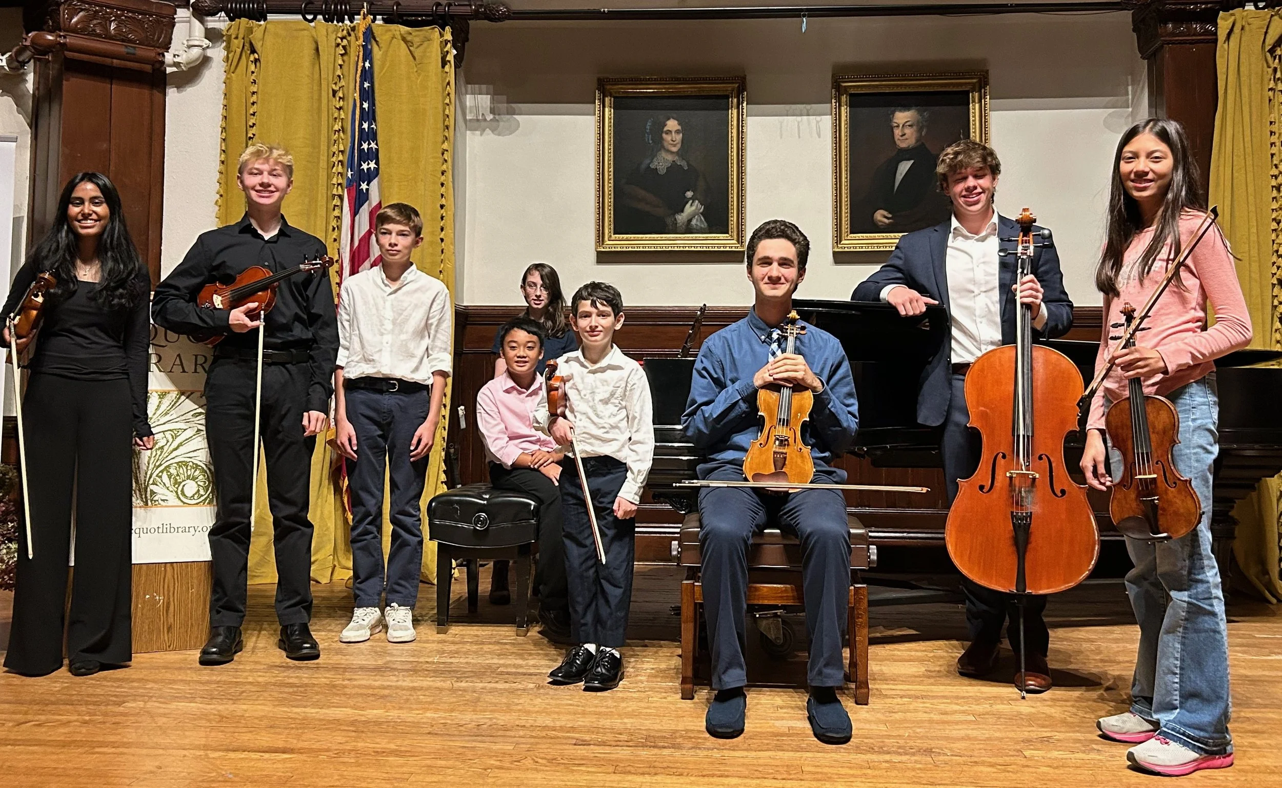 A group of young musicians with string instruments, including violins and cellos, posing for a photo on a stage with a grand piano in the background, in a room with framed portraits and yellow curtains.