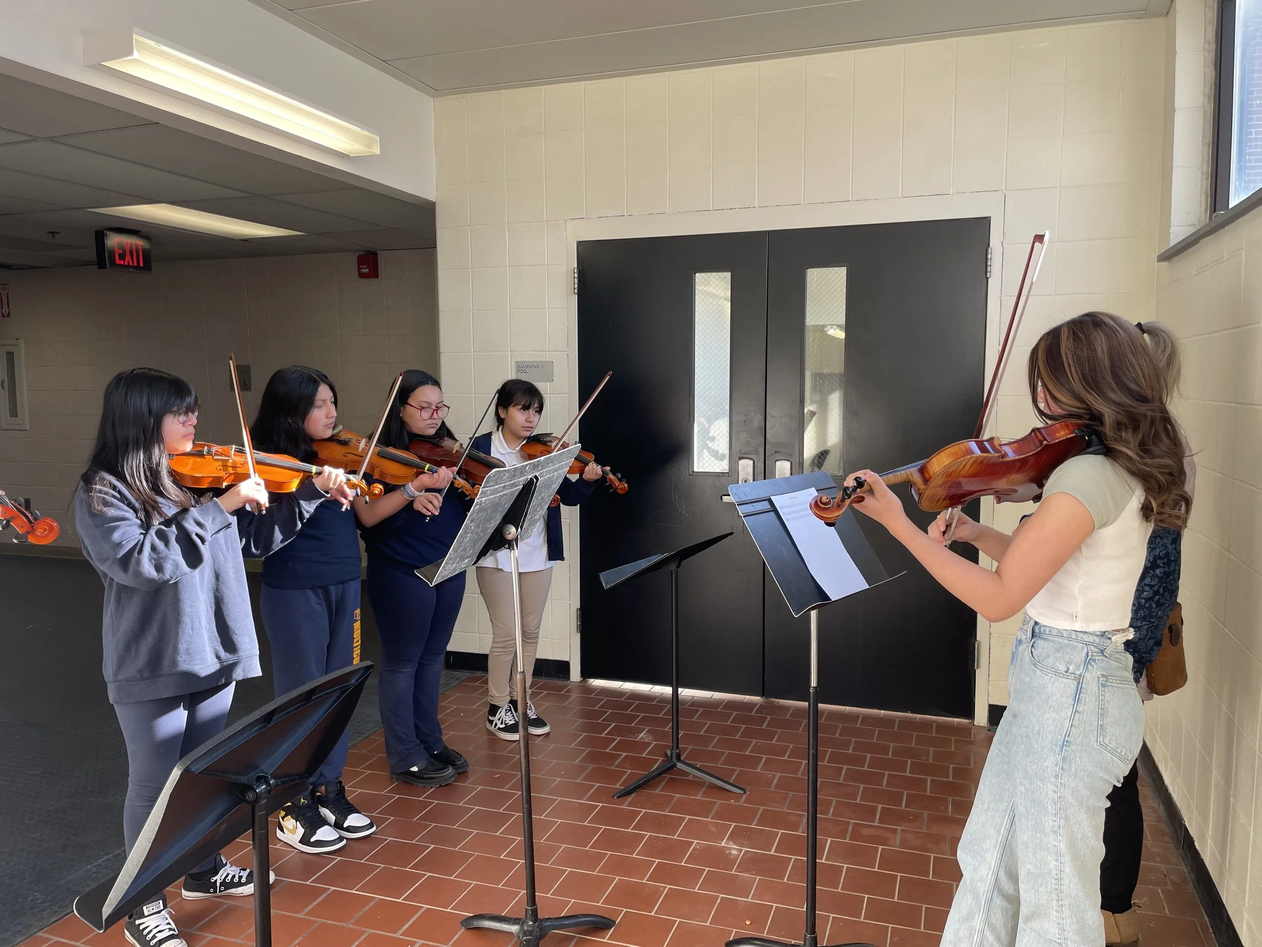 A group of young girls practicing violin in a hallway, focused on their sheet music, with a black door and windows in the background.
