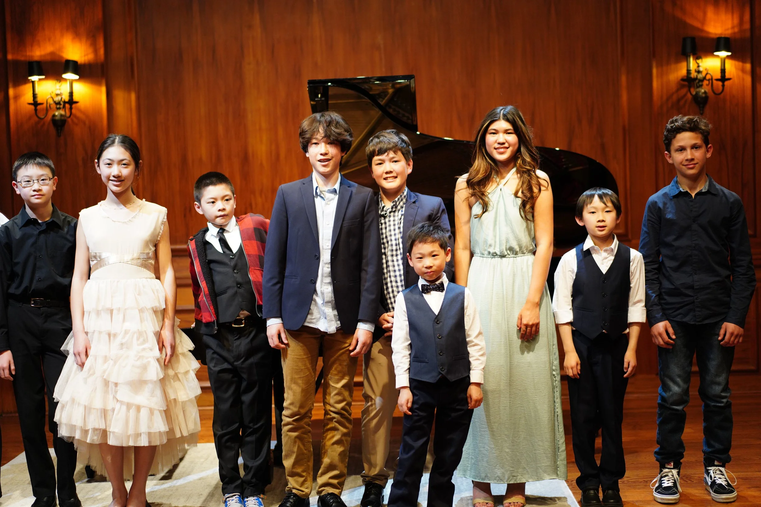 Group of children and a woman standing on a stage in front of a grand piano, with wood-paneled walls and wall sconces lighting, dressed in semi-formal attire for a performance or recital.