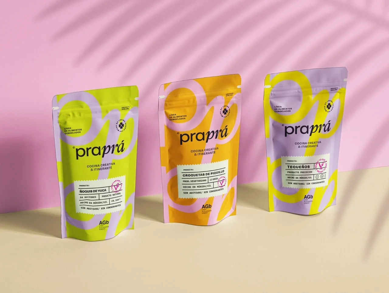 Tres paquetes de alimentos congelados de diferentes sabores en envoltorios de colores pastel, con fondo rosa y sombra de planta