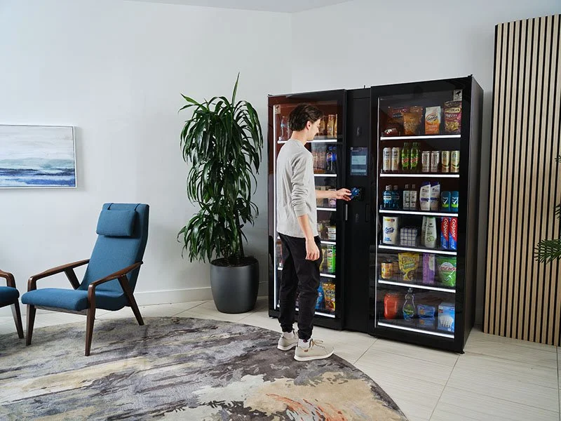 A person using a vending machine in a modern lounge area with a blue chair, a large potted plant, and a colorful abstract rug.