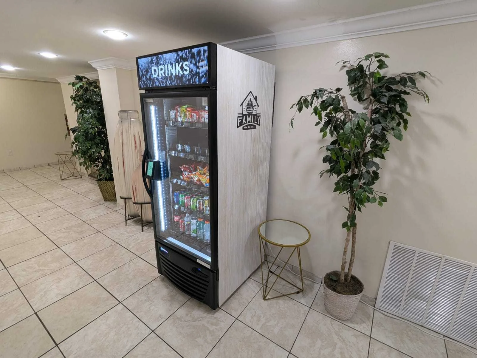 Vending machine with snacks and drinks in a hallway with tiled floor and potted plants.