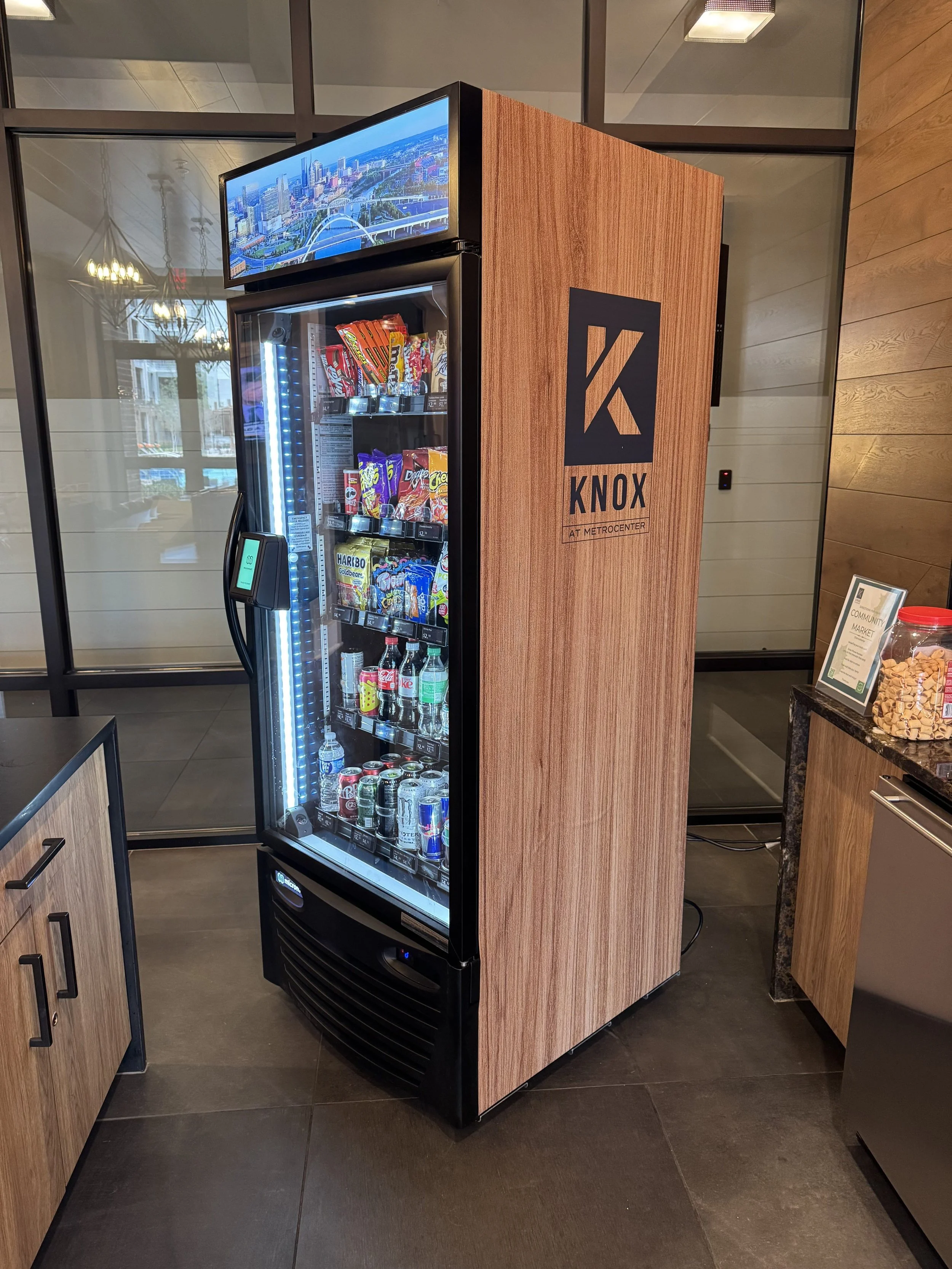 A vending machine in a coffee shop with snack and drink options inside, labeled with 'Knox at Metrocenter', situated next to a wooden counter with snacks and a framed sign.