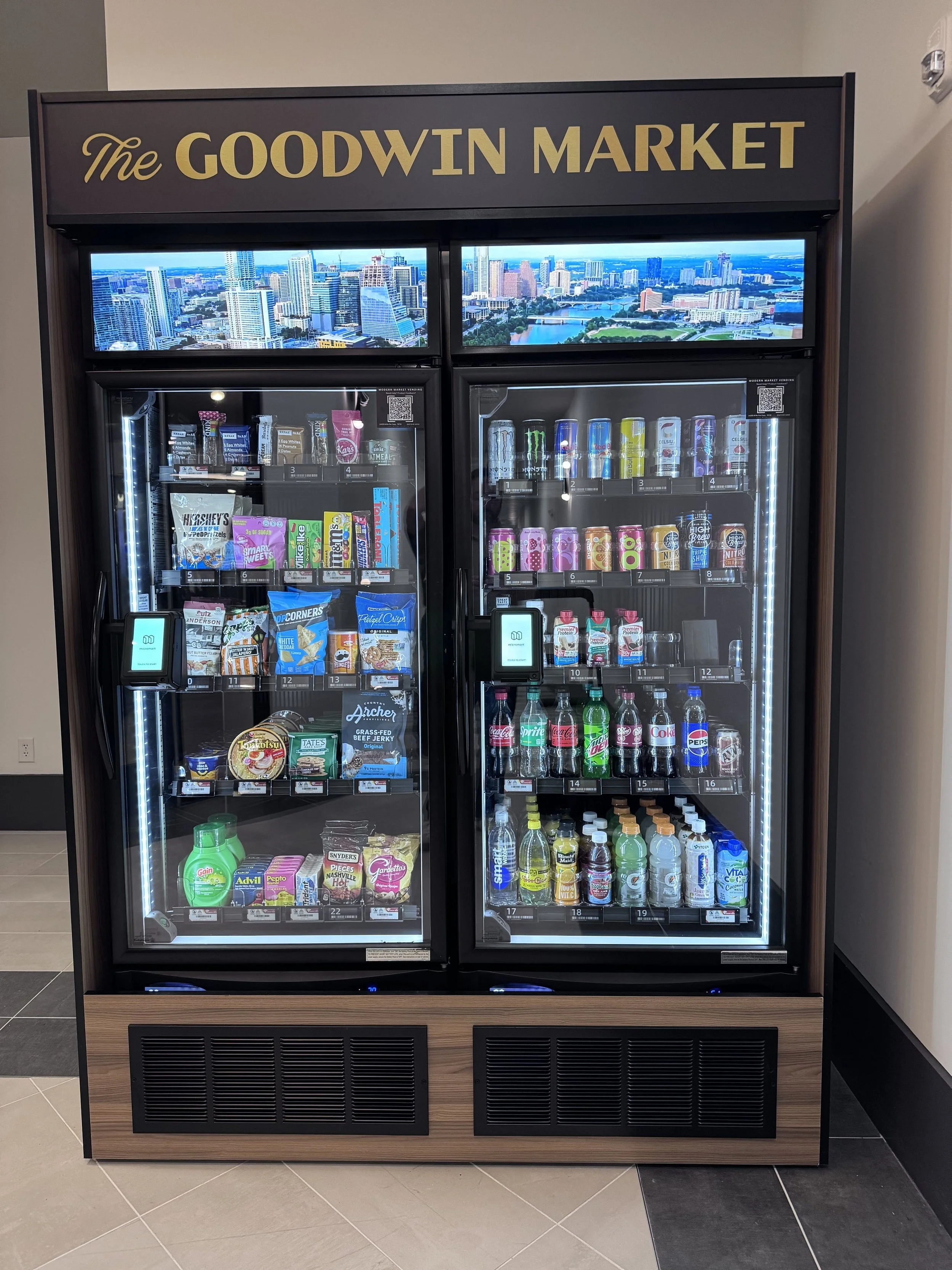Vending machine labeled 'The GOODWIN MARKET' with snacks and beverages inside, situated indoors against a wall, with a cityscape photo at the top.