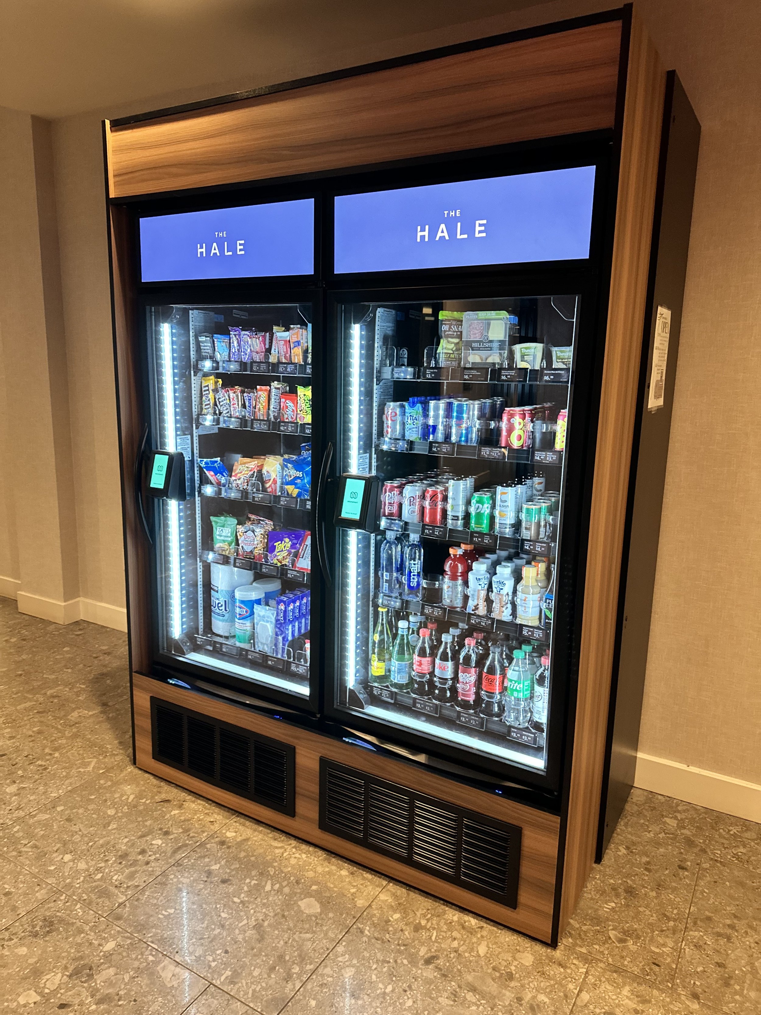 Two refrigerated vending machines with snacks and drinks, labeled 'The Hale,' inside a building with marble tile flooring and neutral-colored walls.