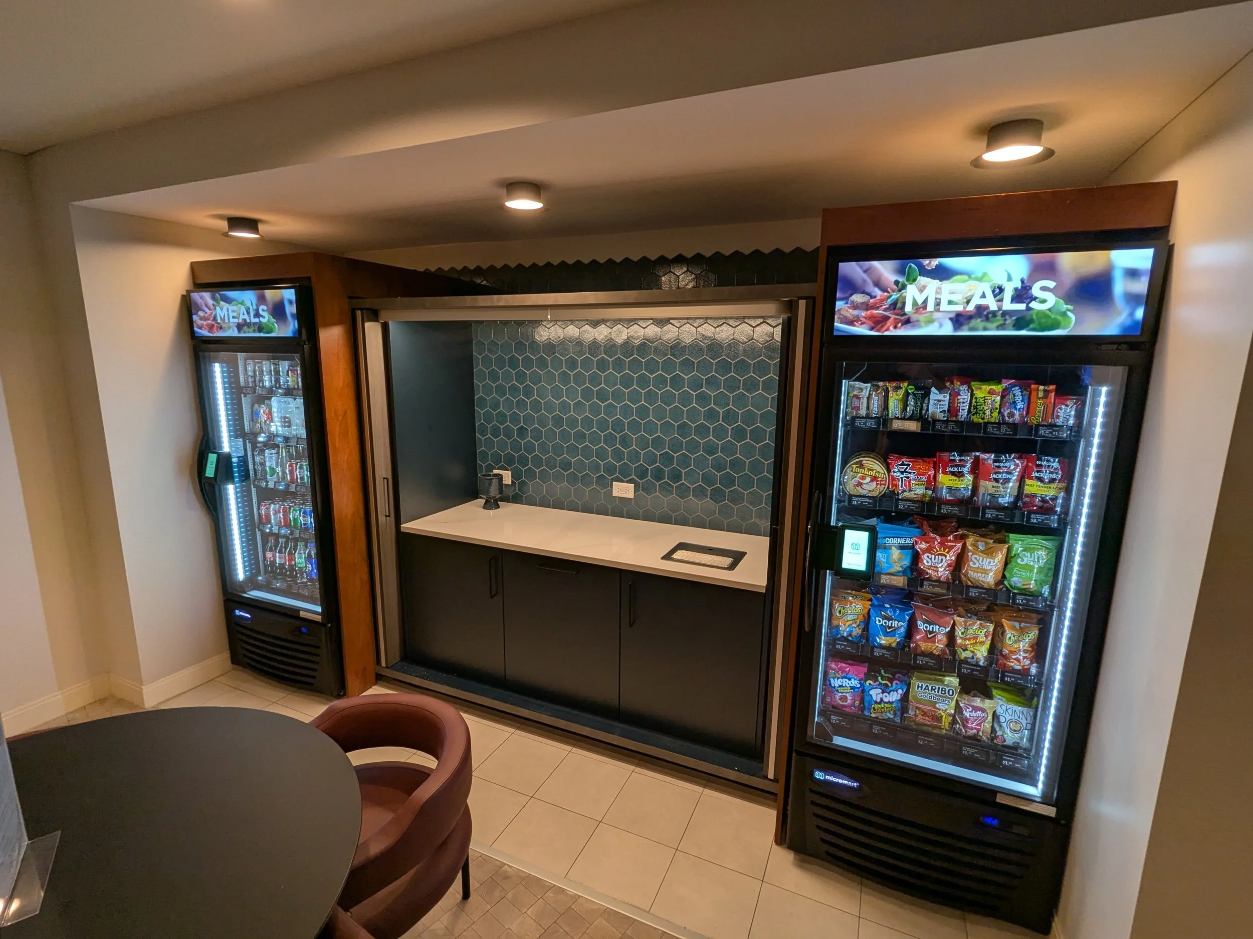 Vending machine filled with snacks on the right, snack storage on the left, and a counter area in the middle with a blue honeycomb backsplash and a sanitizer dispenser. A sign says 'MEALS'.