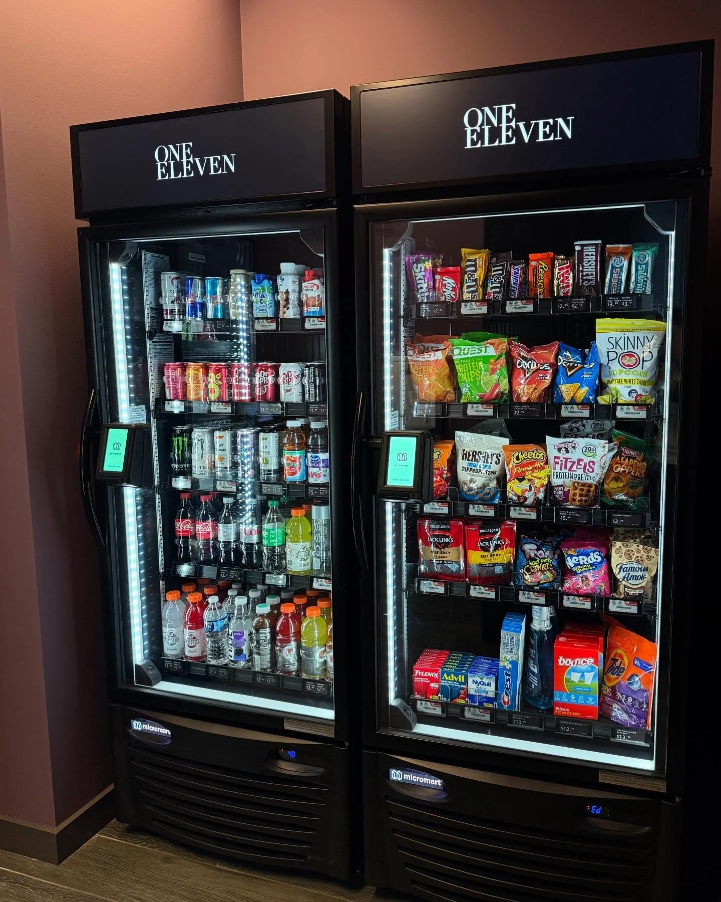 Two vending machines stocked with snacks and beverages, located in a corner against a pink wall.