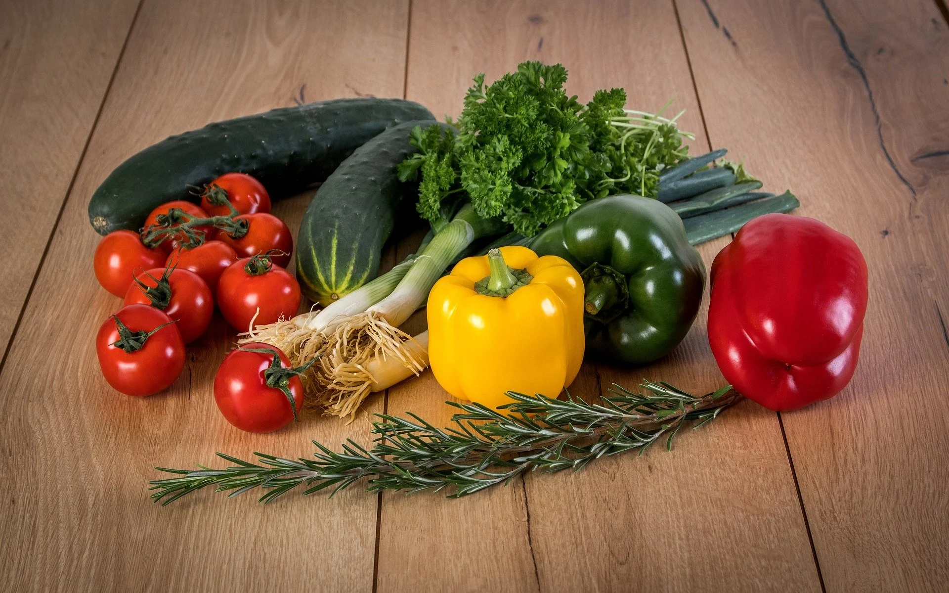 Assorted fresh vegetables on a wooden surface, including cherry tomatoes, cucumbers, green bell pepper, red bell pepper, yellow bell pepper, green onions, parsley, and rosemary.
