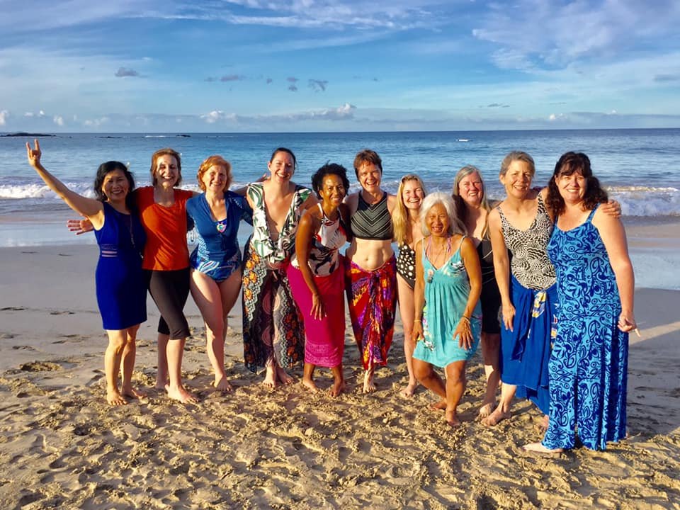 Group of women in colorful dresses and swimsuits standing on a sandy beach with ocean and sky in the background, smiling and posing for a photo.