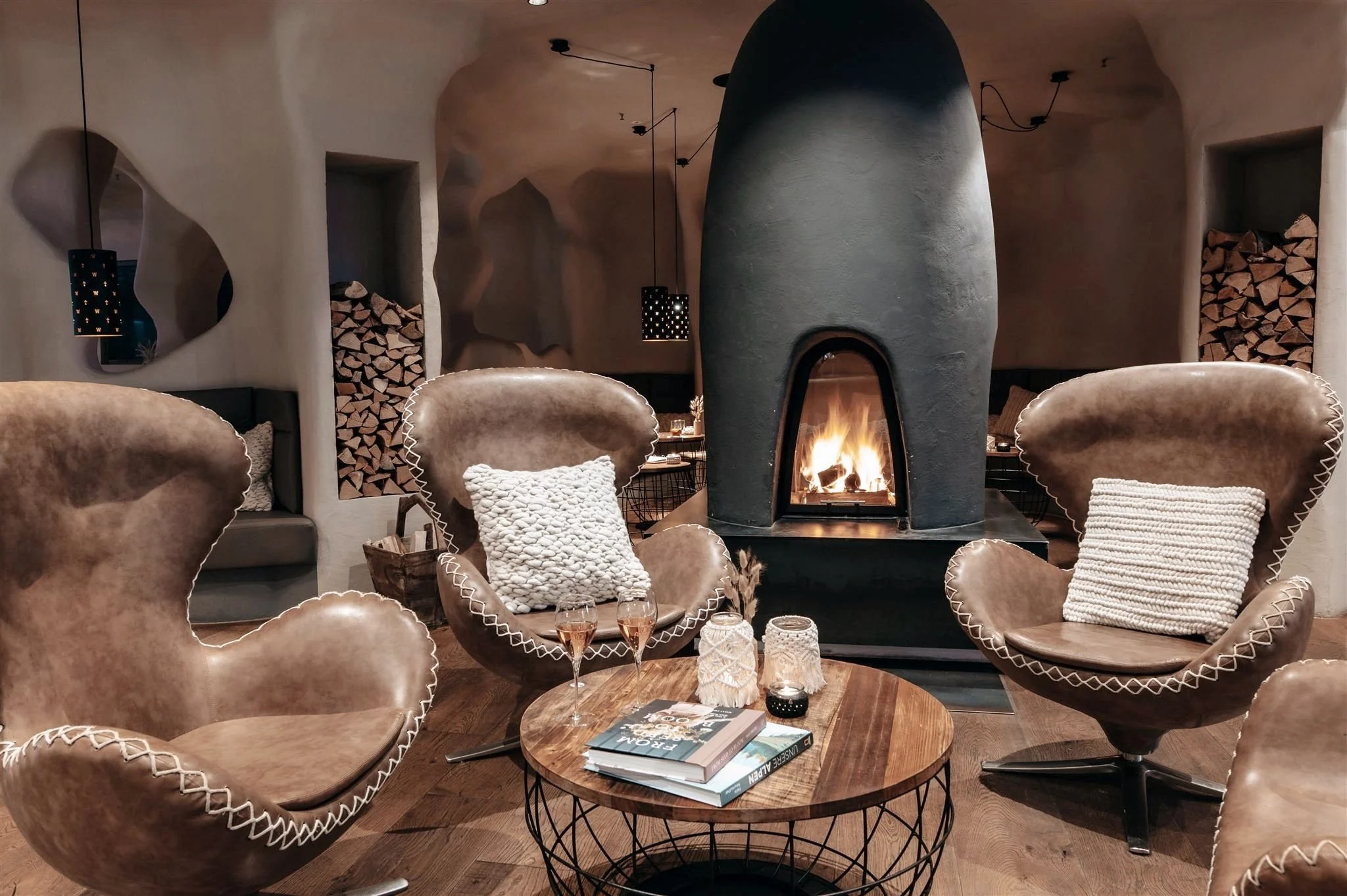 A black fireplace surrounded by cozy leather chairs and freshly cut wood in a cavernous room.
