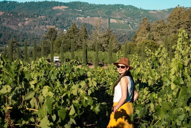 A woman poses in a vineyard, with a sun hat, sunglasses, and a midi skirt.