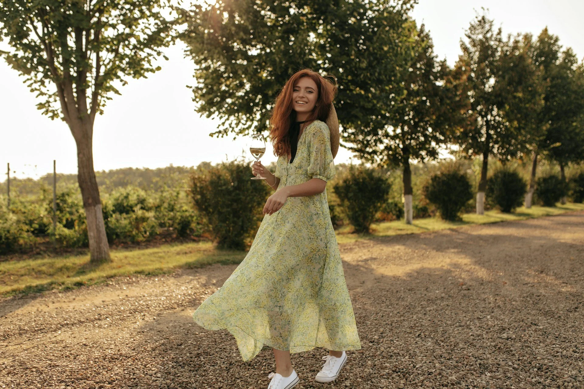 A woman wears a sundress and sneakers in a vineyard, with a glass of wine in hand.