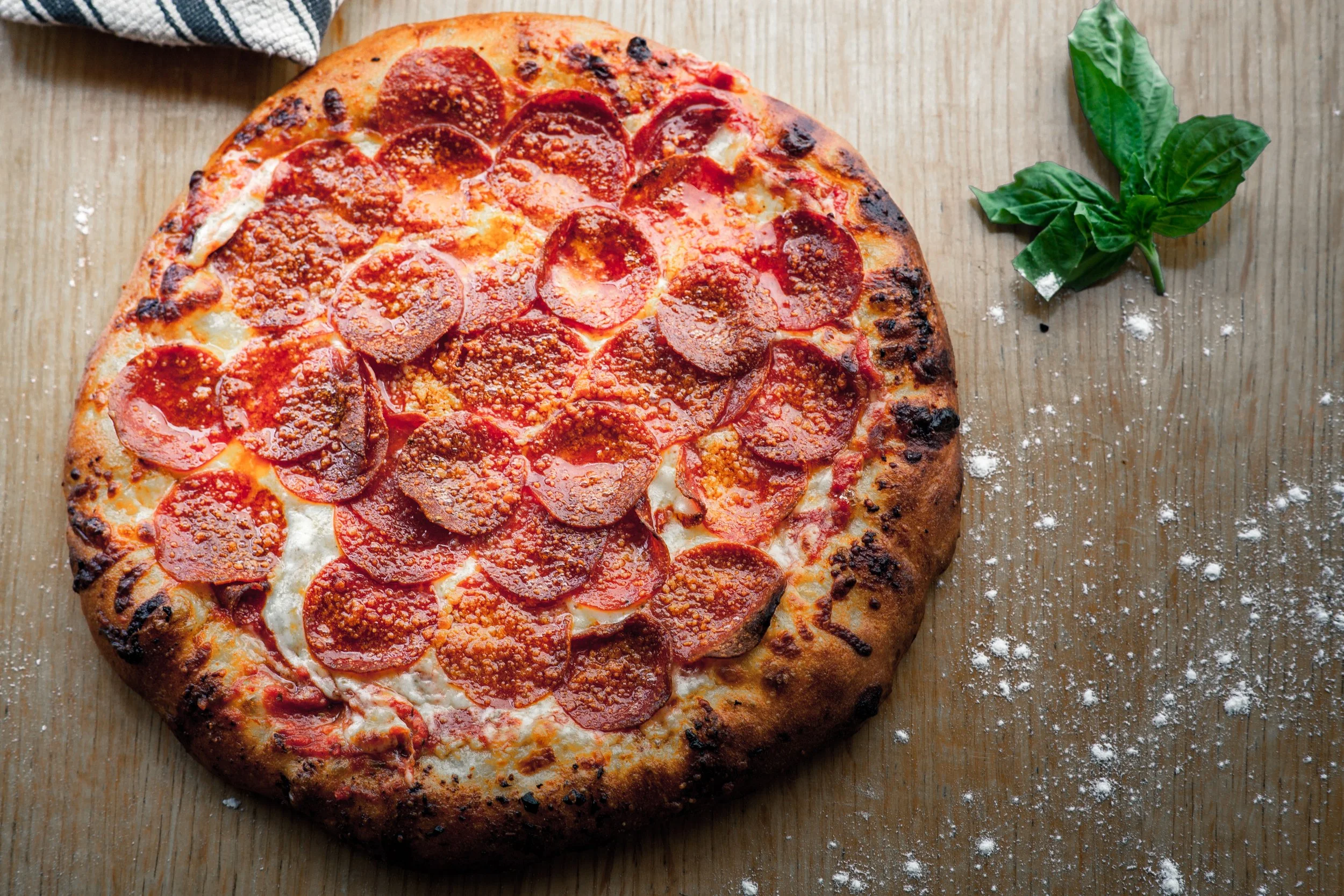 Pepperoni pizza with a golden-brown crust on a wooden surface, sprinkled with some white flour, accompanied by a sprig of fresh basil.