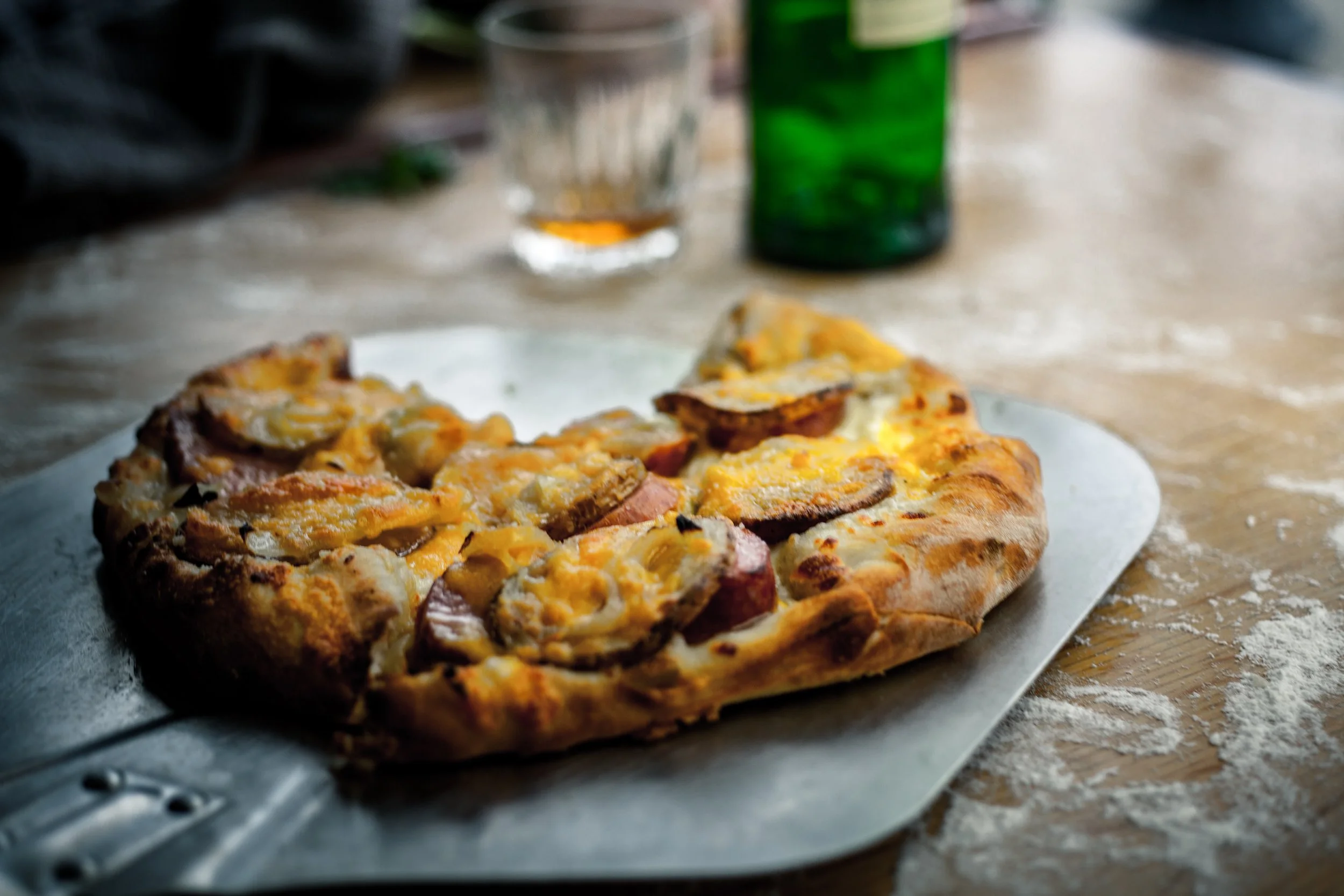Slice of pizza with sausage and cheese on a metal tray, on a wooden table with flour scattered around, with a glass of beverage and a green bottle in the background.