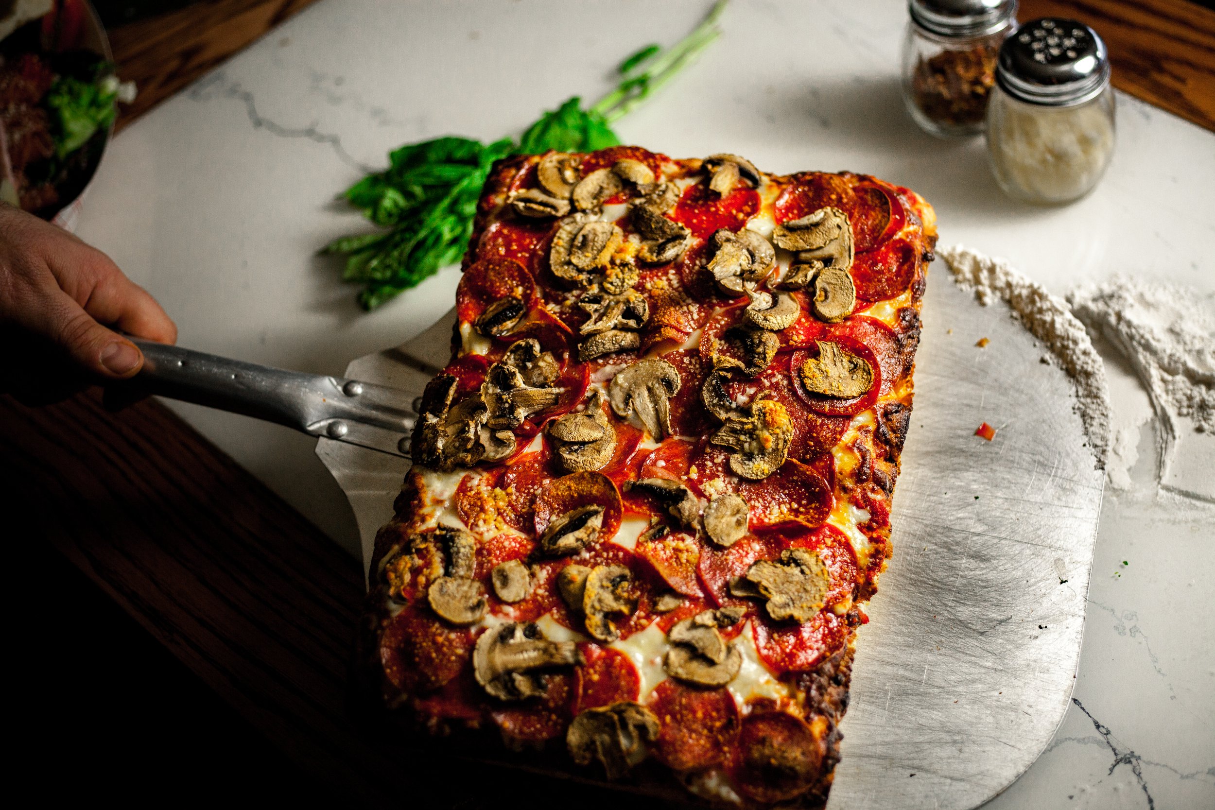 A rectangular pizza with pepperoni, mushrooms, and cheese on a white marble surface. There are green leafy herbs, flour, and spice jars in the background.
