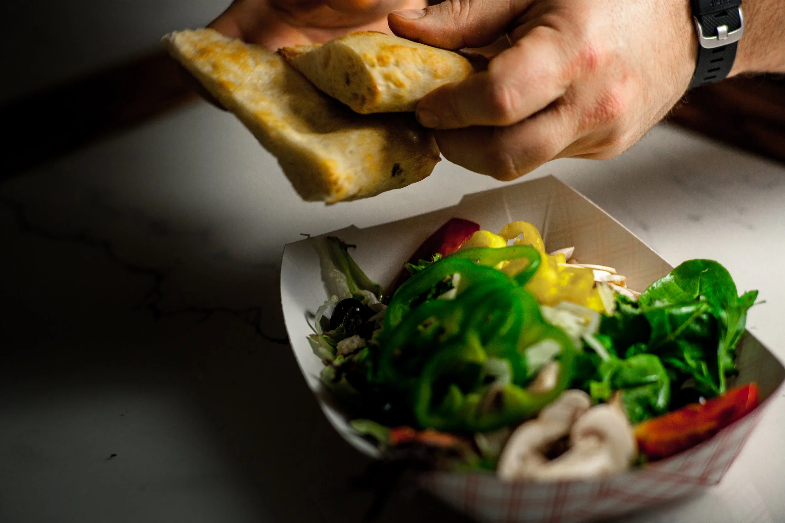 Person holding a piece of pita bread over a box of salad with chopped vegetables, including green bell peppers, mushrooms, and tomatoes.