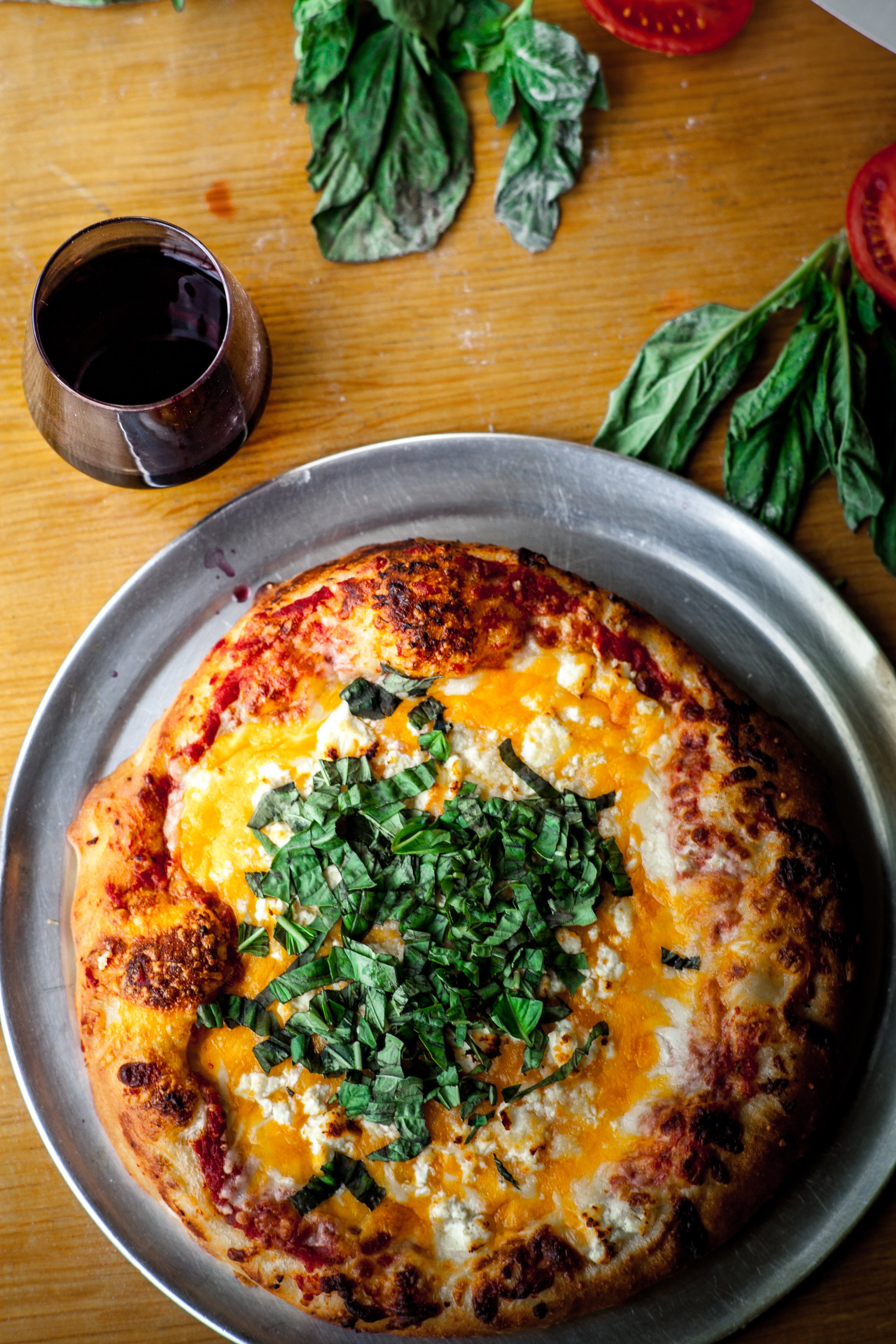 Cheese pizza with chopped basil on top, a glass of red wine, and fresh basil and tomato slices on a wooden table.