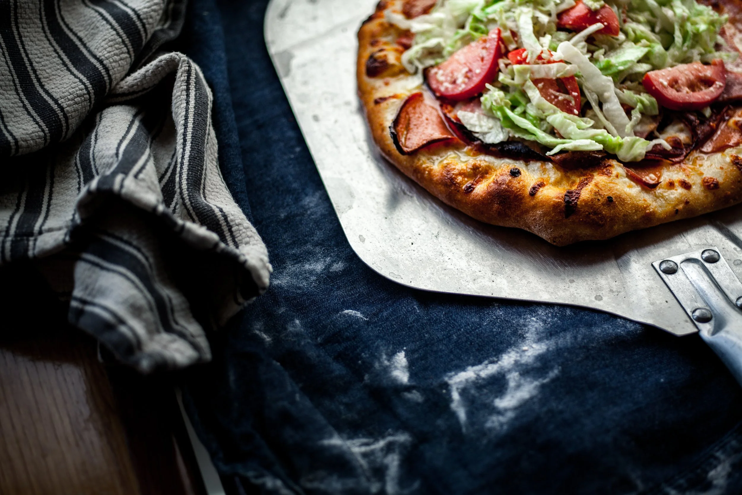 Close-up of a freshly baked pizza with pepperoni, bacon, lettuce, and tomato on top, on a metal pizza peel, with a striped kitchen towel and black cloth on a wooden surface.