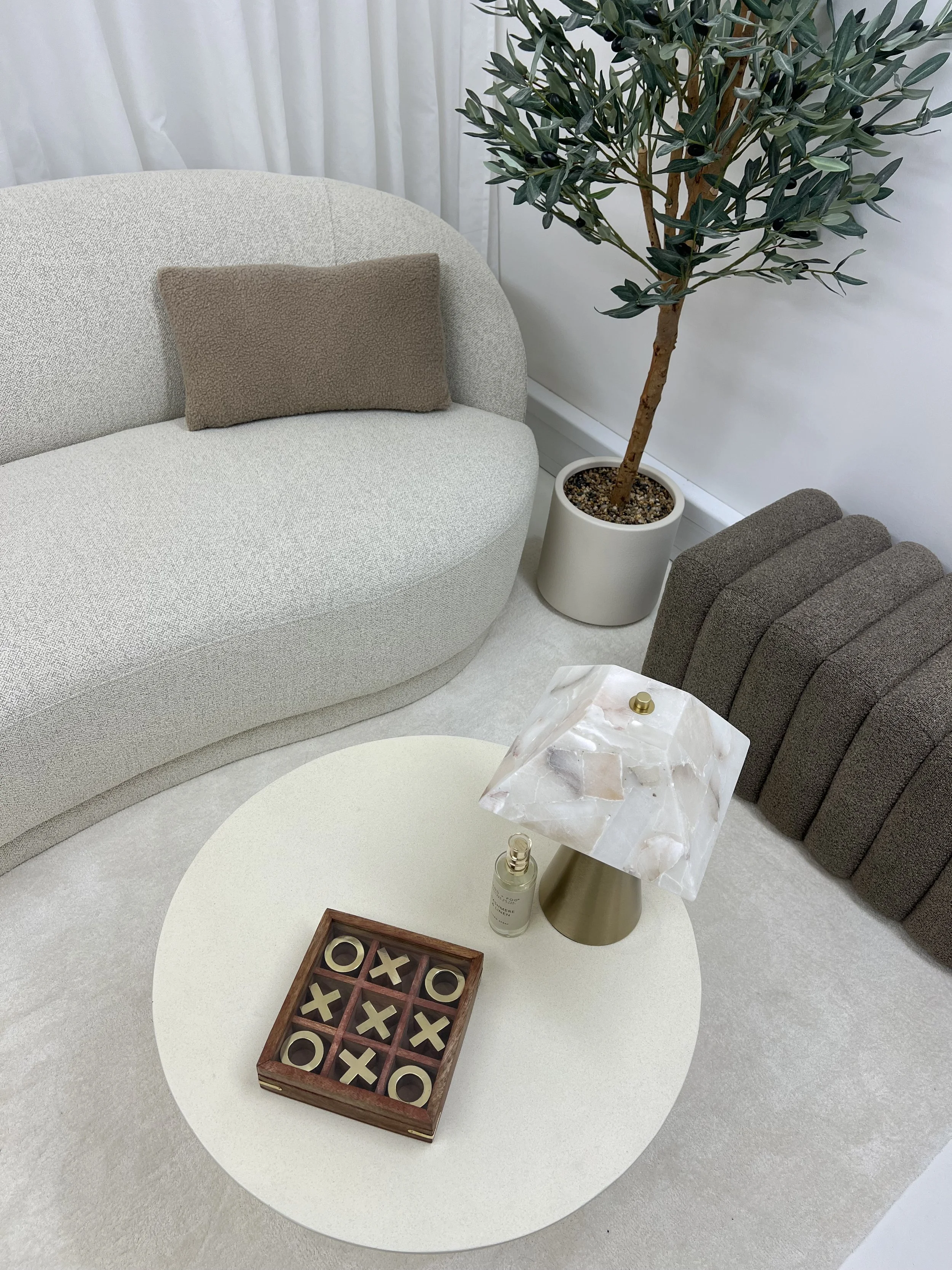 A cozy living room corner with a light beige curved sofa, a brown pillow, a round white coffee table, a decorative marble lamp, a small spray bottle, a tic-tac-toe game, a large potted tree, and a dark gray upholstered cushion or stool on a light-colored carpet.