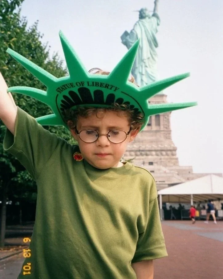 Child Josh Arnon in green and raising his arm like the Statue of Liberty while wearing a green crown in front of the Statue of Liberty.