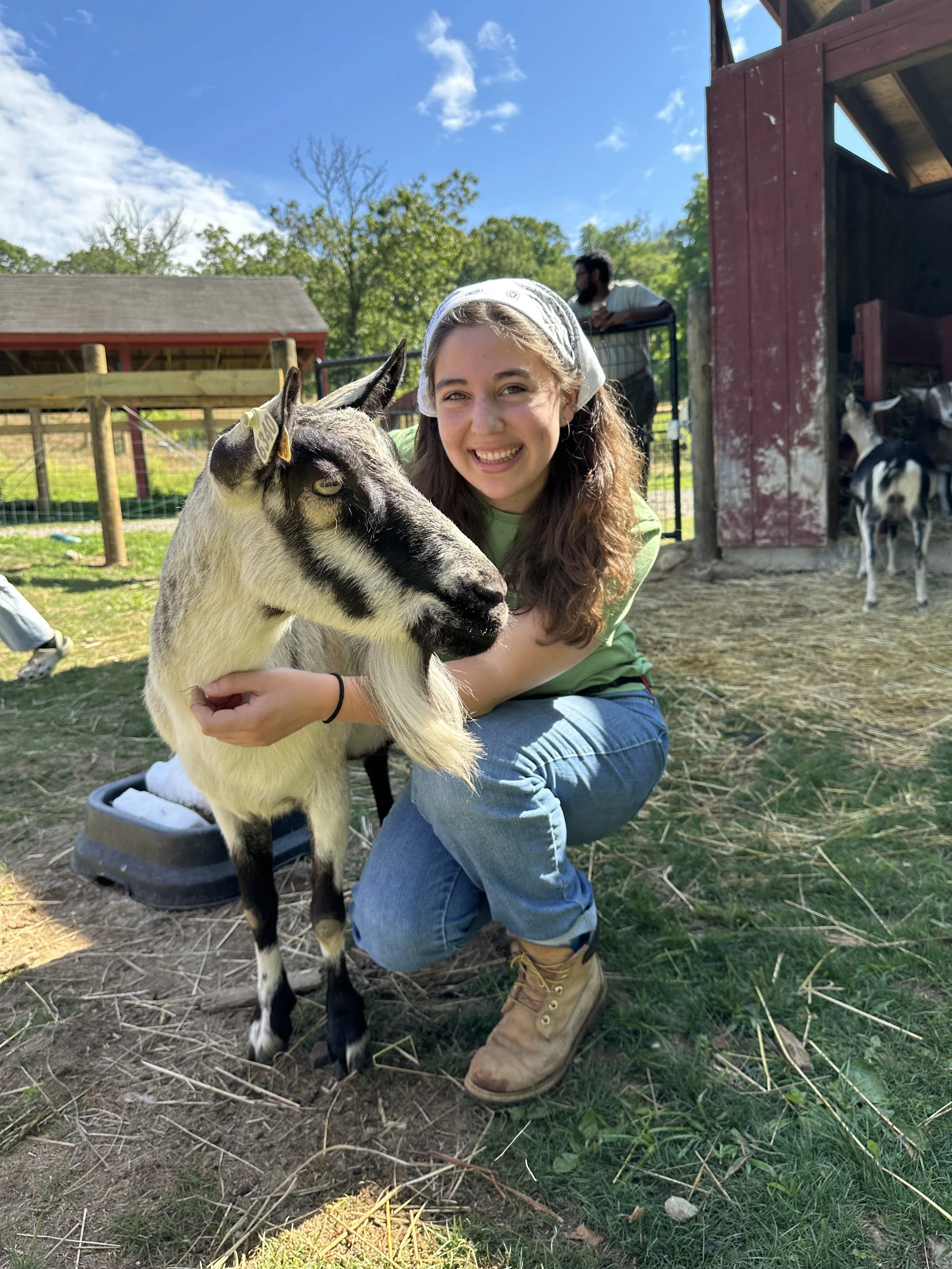 A woman smiling and crouching down next to a baby goat outside on a farm, with a barn and another person in the background.
