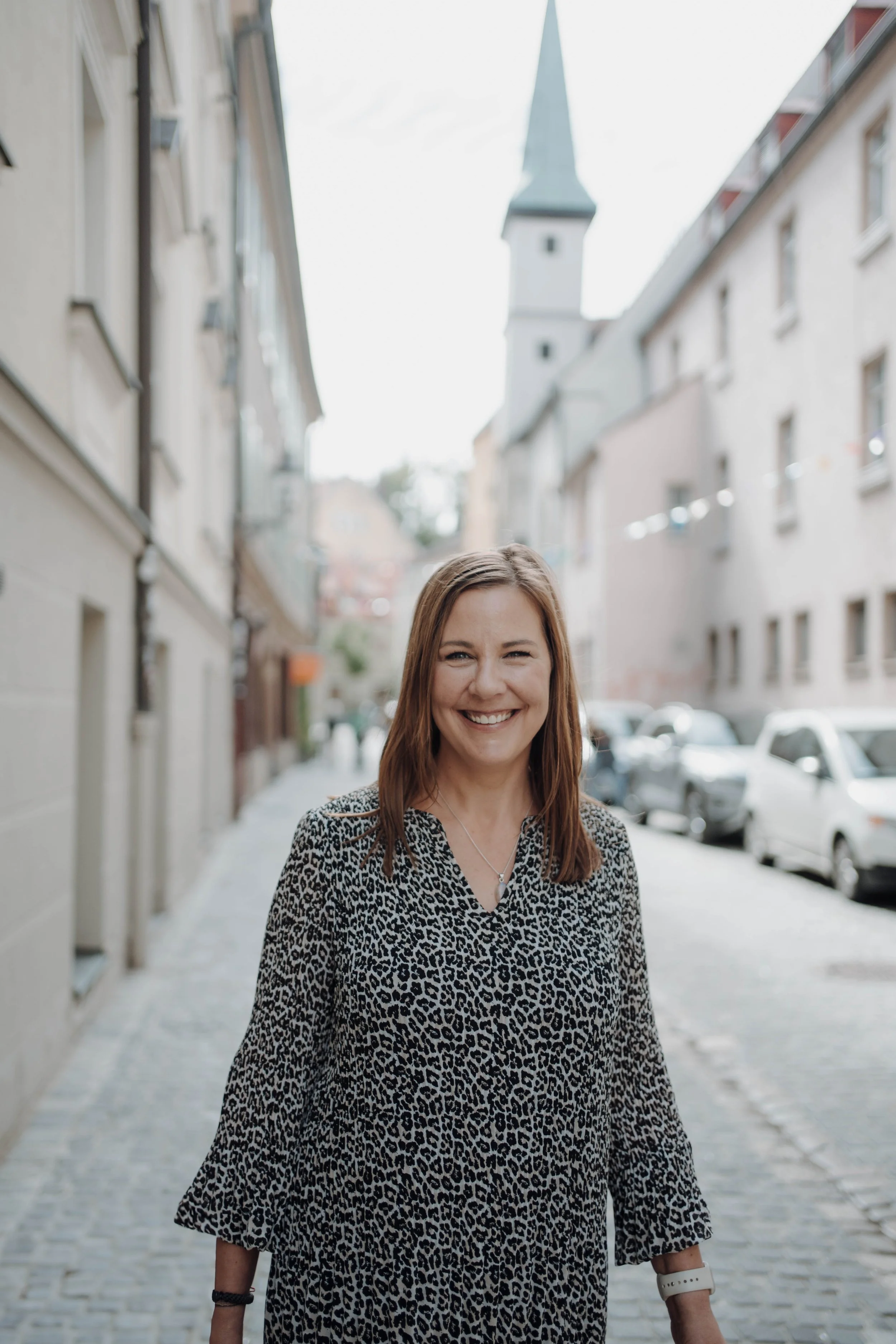 Eine lächelnde Frau mit braunen Haaren, die eine Leoparden-Print-Bluse trägt, steht auf einer Fußgängerstraße in einer Stadt mit alten Gebäuden und einem Kirchturm im Hintergrund.