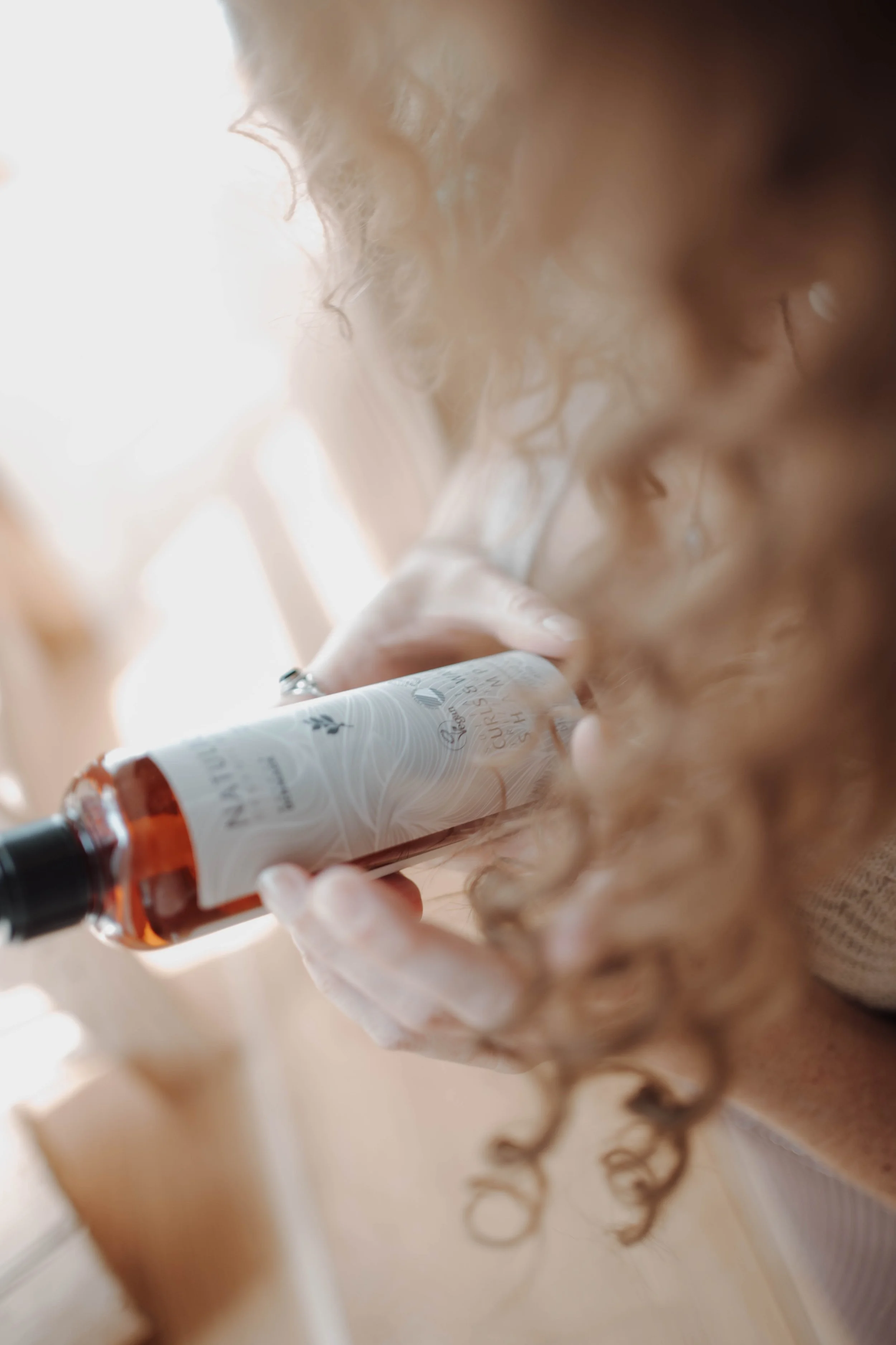 Frau mit lockigem roten Haar trinkt aus einer Flasche, die vermutlich Wein oder Spirituosen enthält, im Raum mit natürlichem Licht.