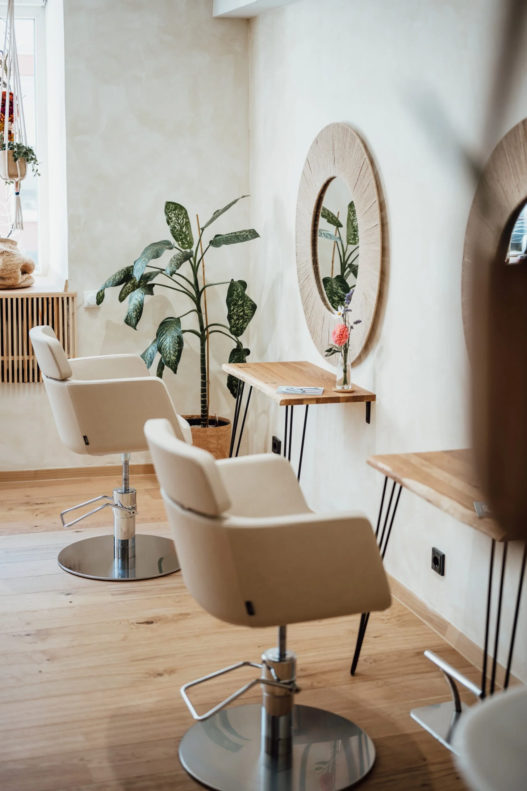 Innenraum eines Friseursalons mit drei weißen Friseurstühlen, einem großen Pflanze, zwei Wandspiegeln und kleinen Holztischen