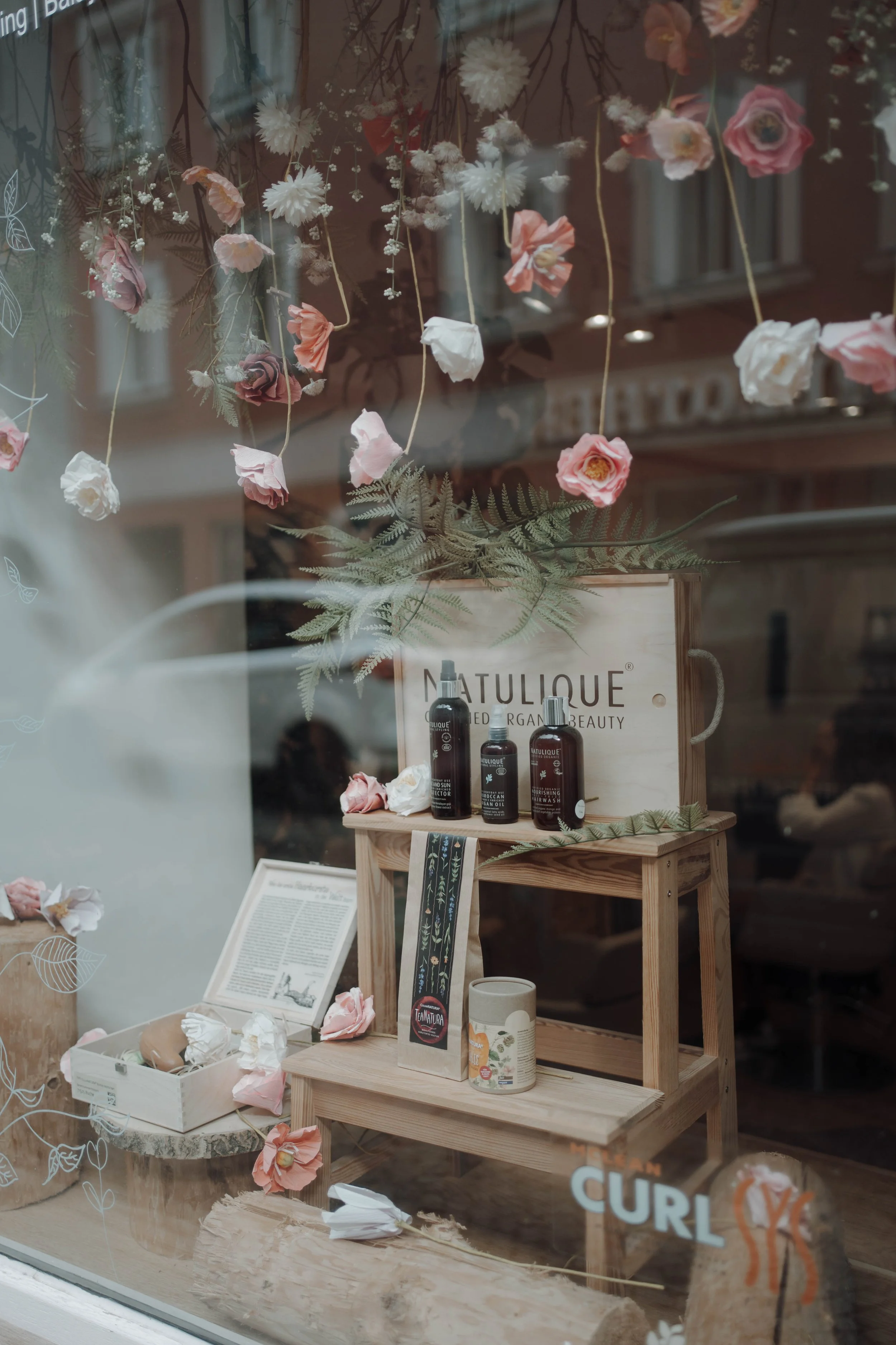 Fensterdekoration mit hängenden Papierblumen in Rosa, Weiß und Pfirsichfarben. Im Inneren ein Holztisch mit Schönheitsprodukten, Pflanzen und Dekorationsartikeln.