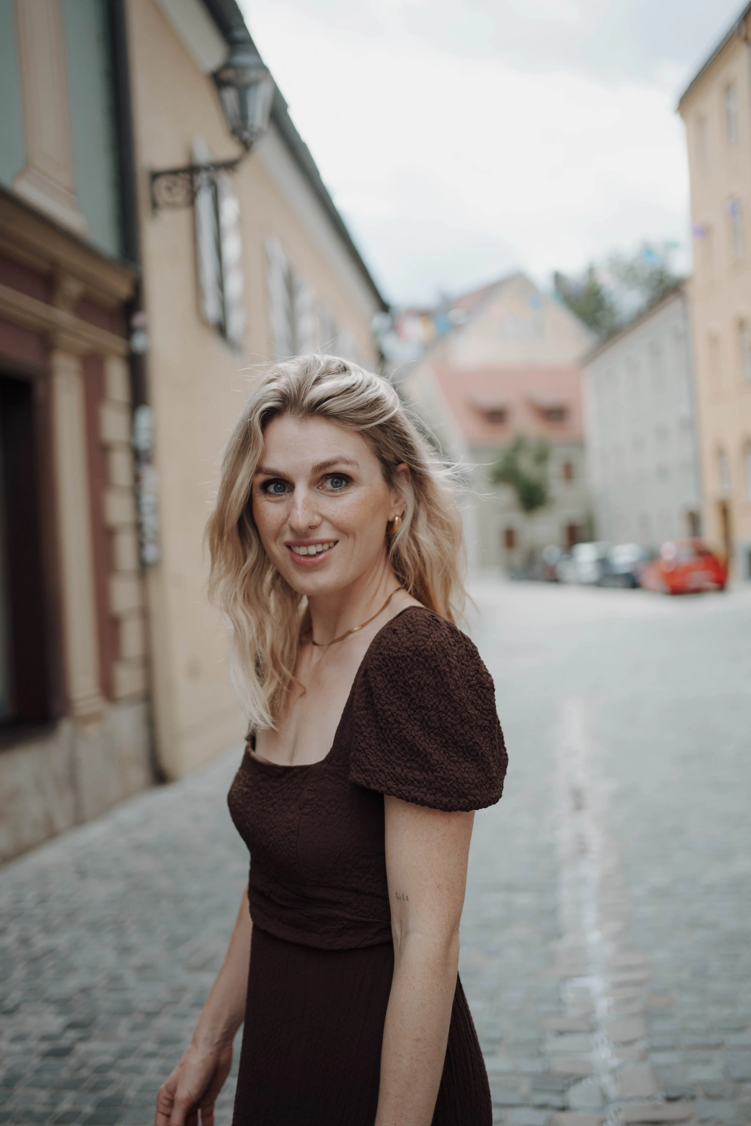Eine blonde Frau in einem dunklen Kleid steht auf einer gepflasterten Straße in einer europäischen Altstadt, im Hintergrund sind alte Gebäude sichtbar.