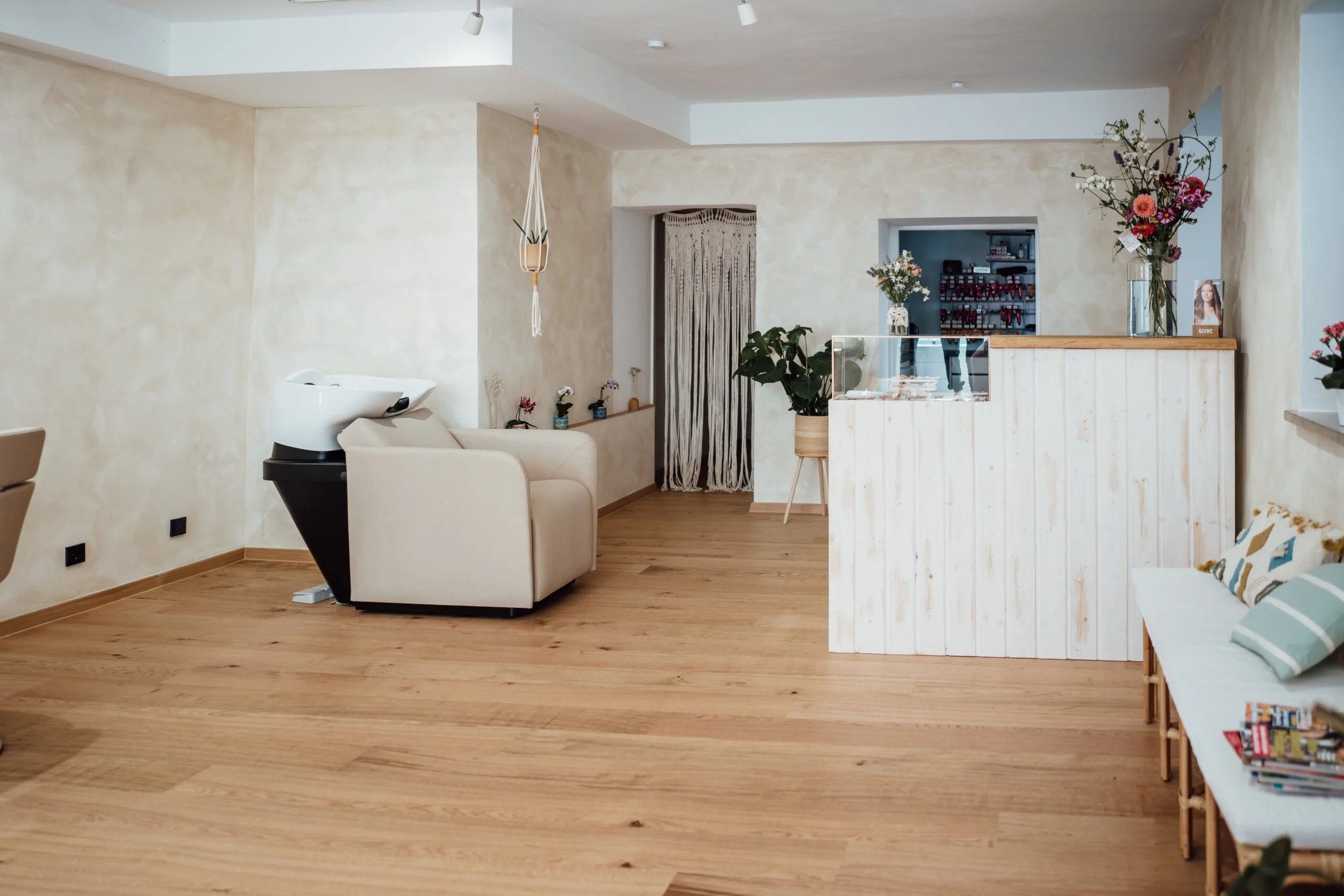 Innenansicht eines stilvollen, hellen Raumes mit Holzfußboden, weißen Wänden und gemütlichen Möbeln, darunter ein Sofa mit Kissen, eine Bar mit Blumenvasen, Pflanzen und Dekorationen.