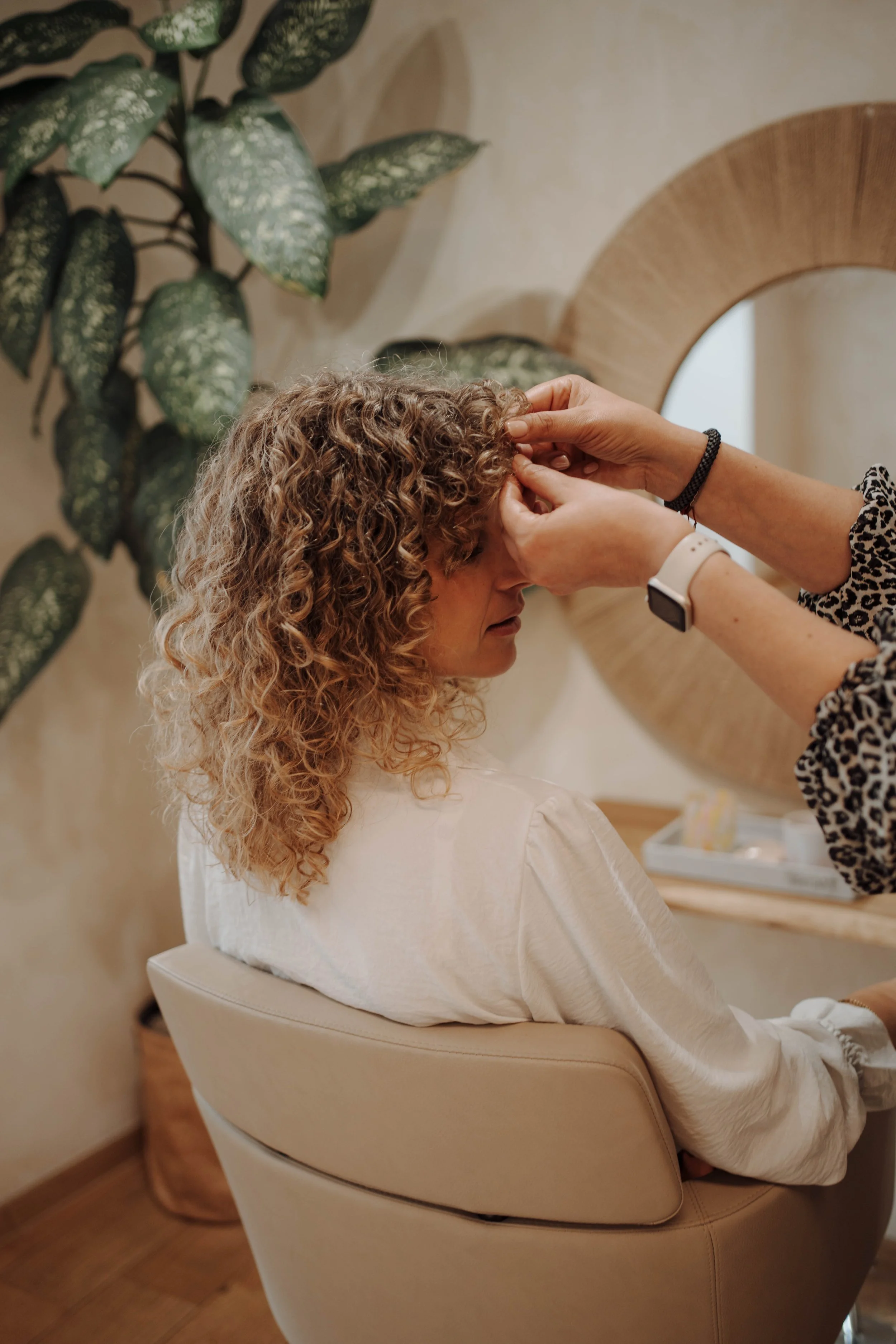 Eine Frau mit lockigem Haar sitzt in einem Friseurstuhl, während eine Friseurin an ihrem Haar arbeitet. Im Hintergrund sind Pflanzen und eine runde Holzdeko zu sehen.