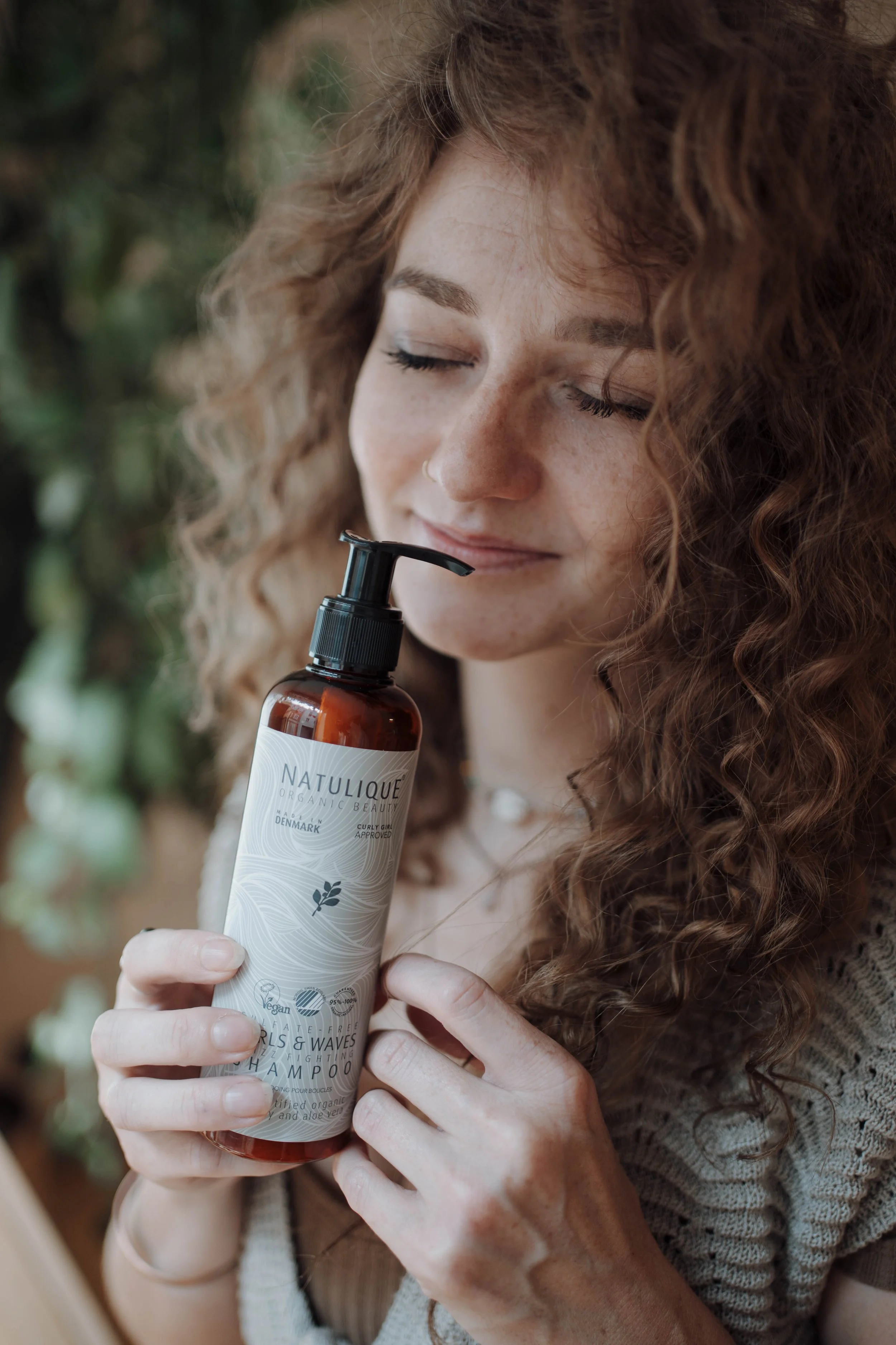 Eine junge Frau mit lockigem, rotem Haar hält eine Flasche Shampoo mit Pumpverschluss und lächelt leicht, mit geschlossenen Augen, vor grünem, unscharfem Hintergrund.