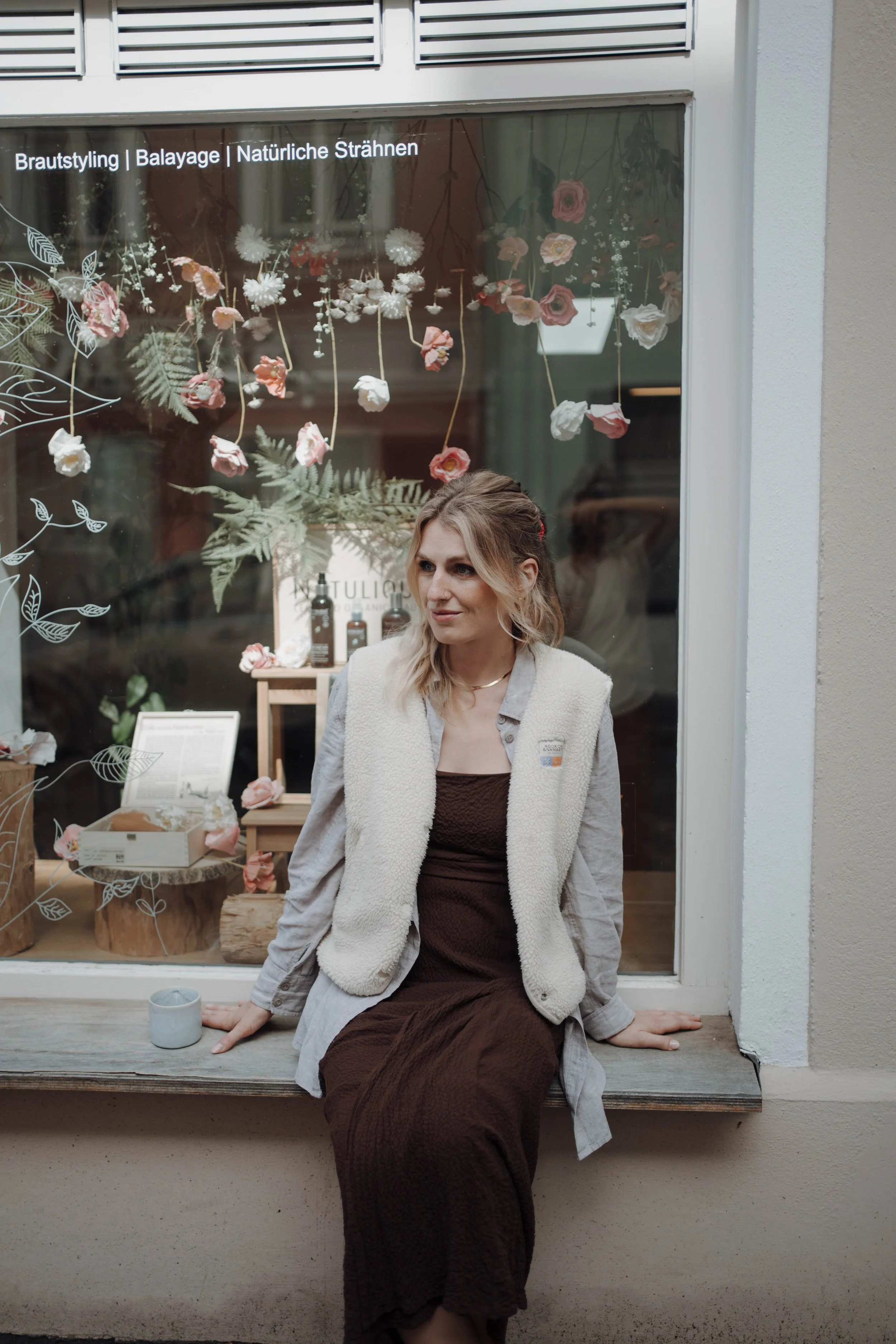 Eine Frau sitzt auf einem Fensterbank vor einem Schaufenster mit Blumen- und Pflanzenmotiven. Im Schaufenster sind auch Produkte und Dekorationsartikel zu sehen.